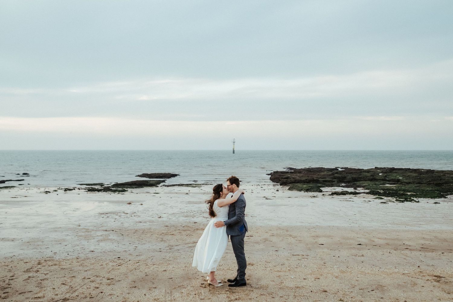 Wedding couple embracing on rocky beach during overcast day with ocean backdrop.