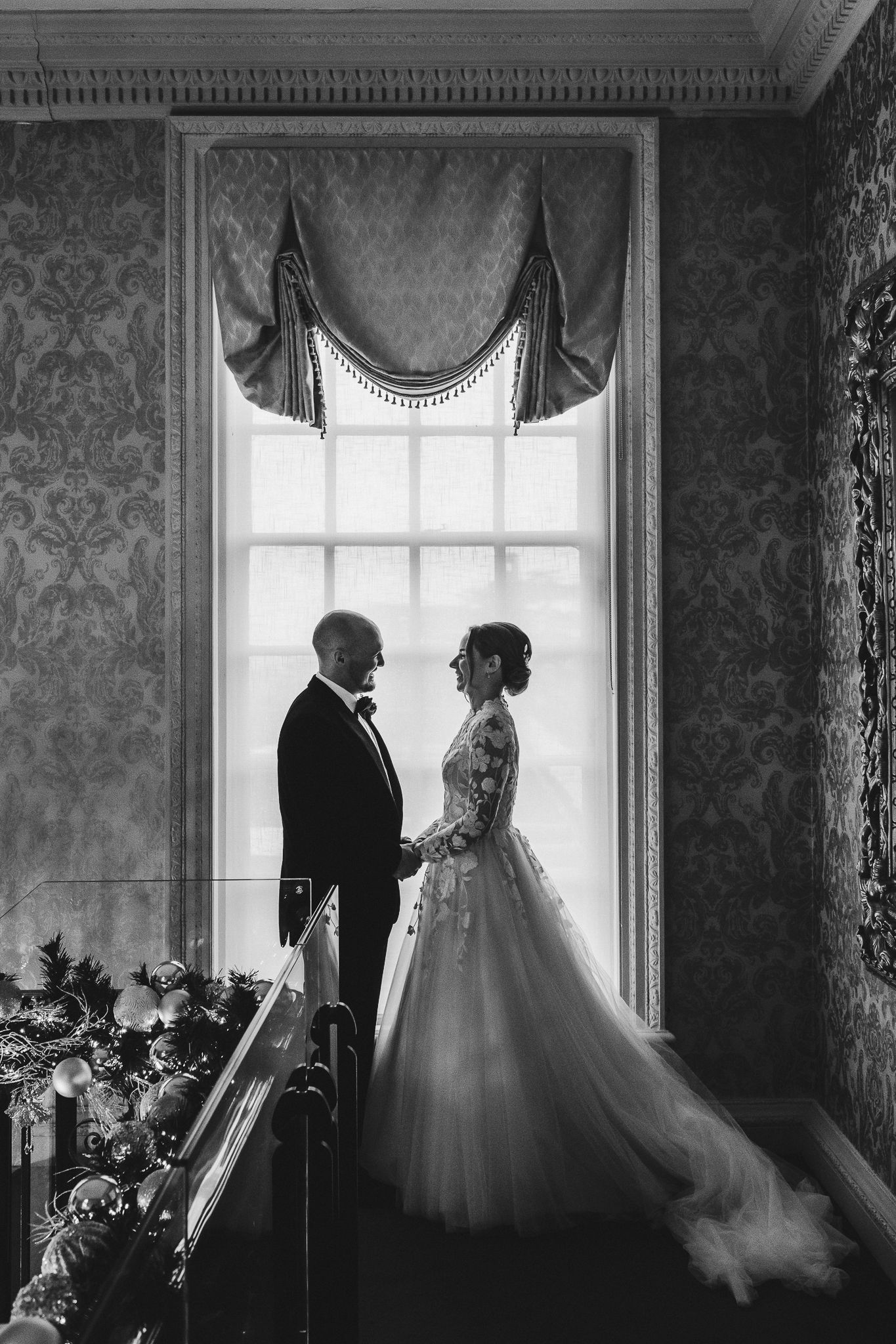 Silhouetted couple stands by ornate window in vintage-style wedding portrait.
