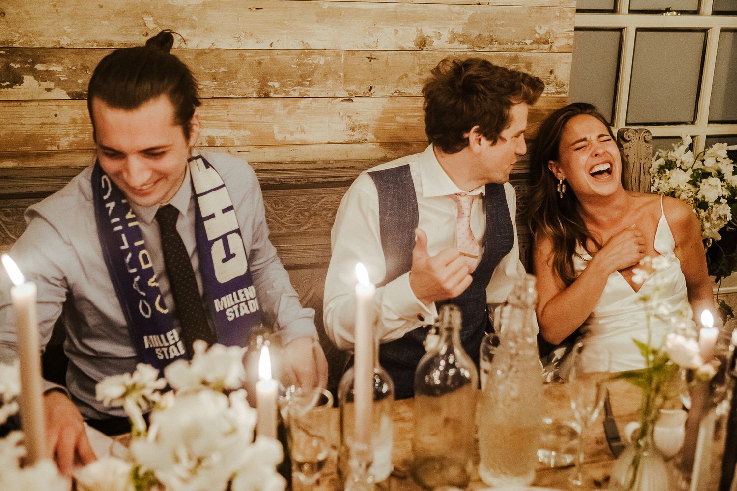 Party guests share genuine laughter at rustic wooden table during dinner celebration.