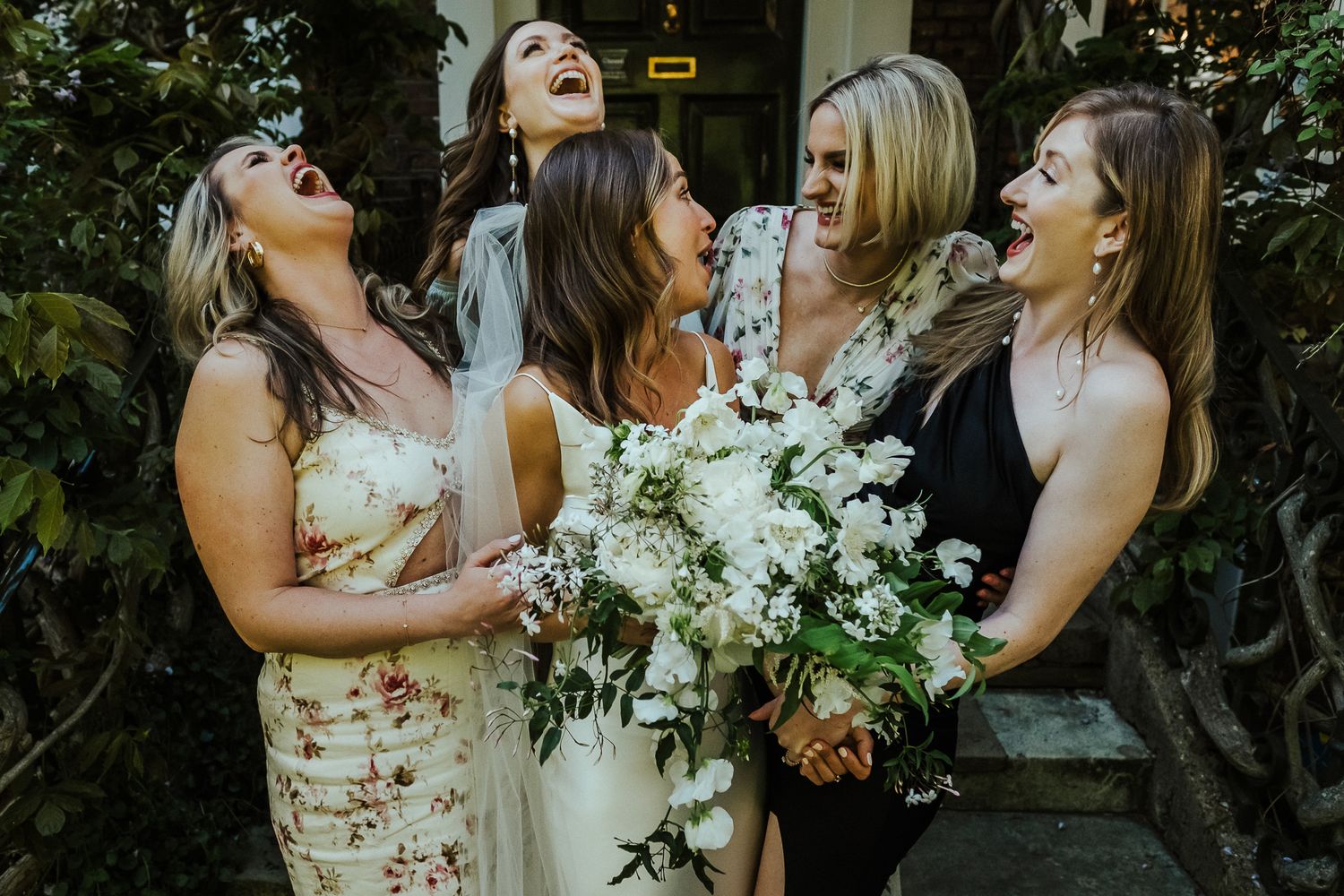 Bridesmaids in floral dresses share laughter while holding white bouquets.