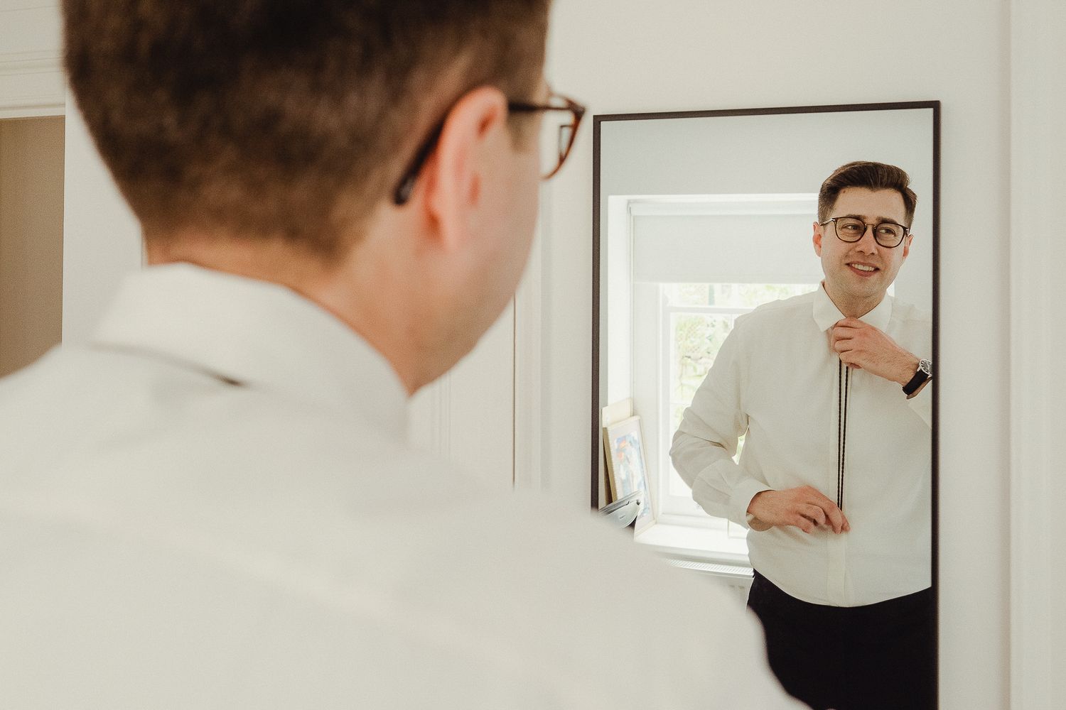 A person wearing glasses and a white shirt looks at their reflection in a bathroom mirror.