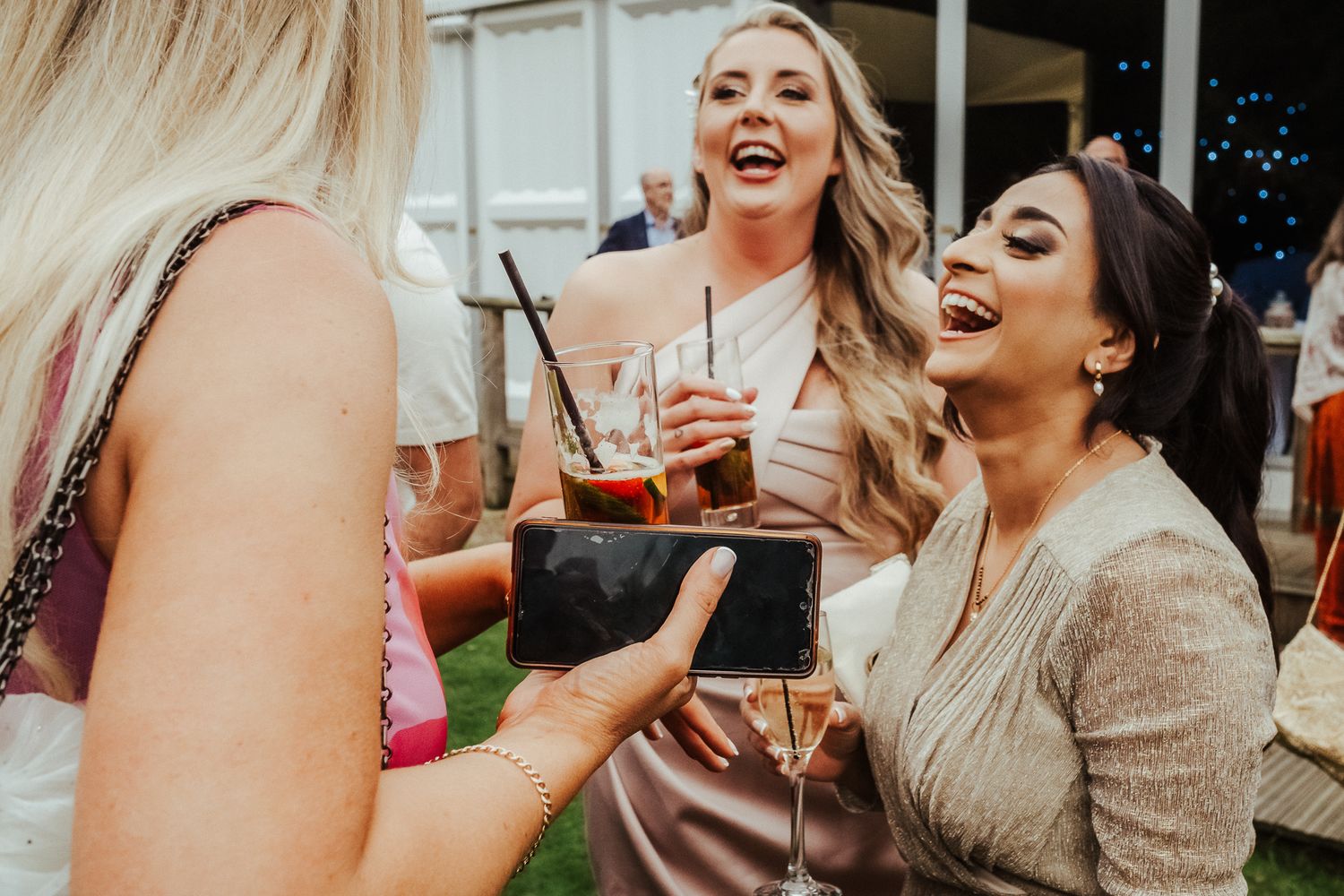 Three friends laugh and share drinks together at an outdoor celebration event in warm sunlight.