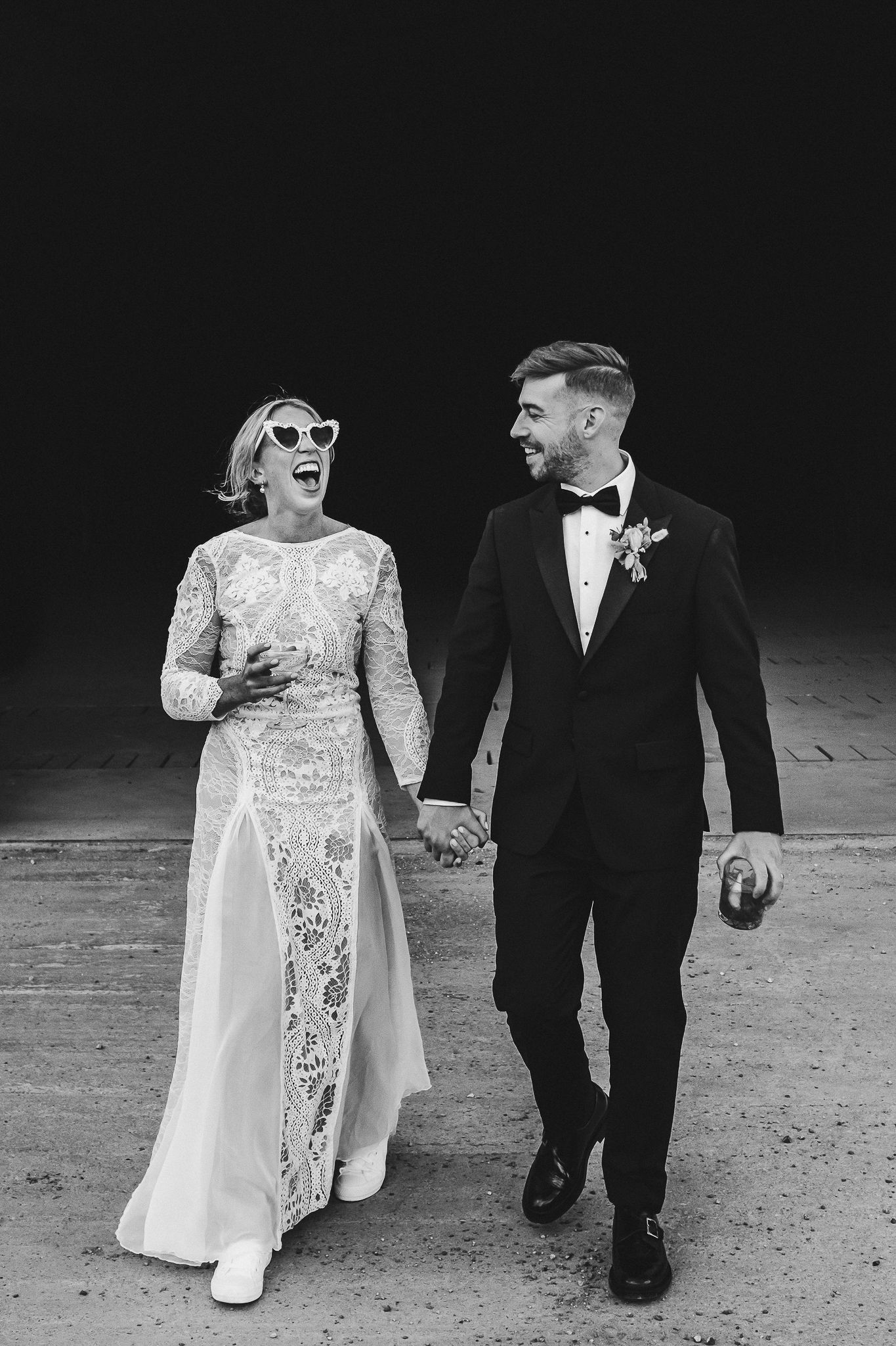 A black and white wedding photo of a couple walking together, with the bride wearing a lace gown and sunglasses.