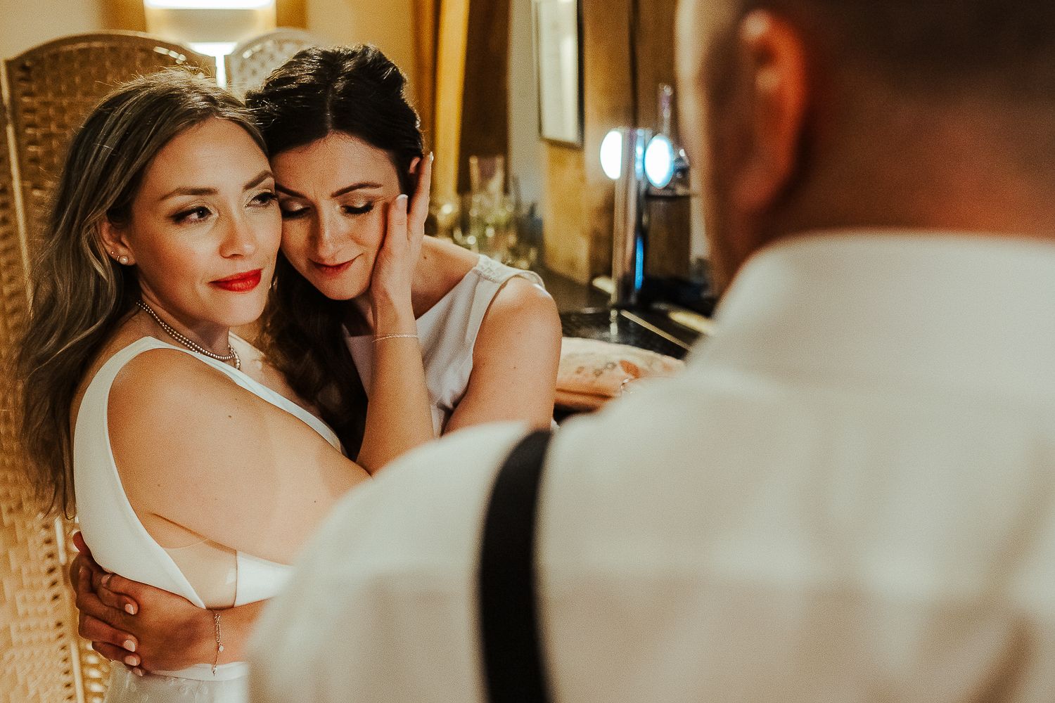 Two women share an intimate moment while getting ready in front of a vanity mirror.