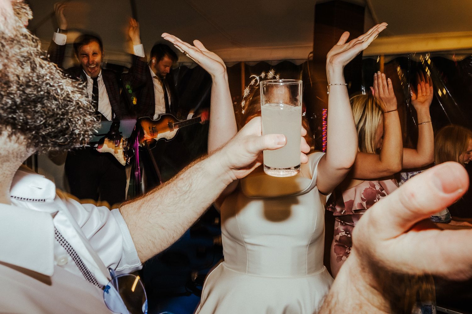 Guests hold up their hands in celebration as the bride holds a wedding cake at a lively reception party.