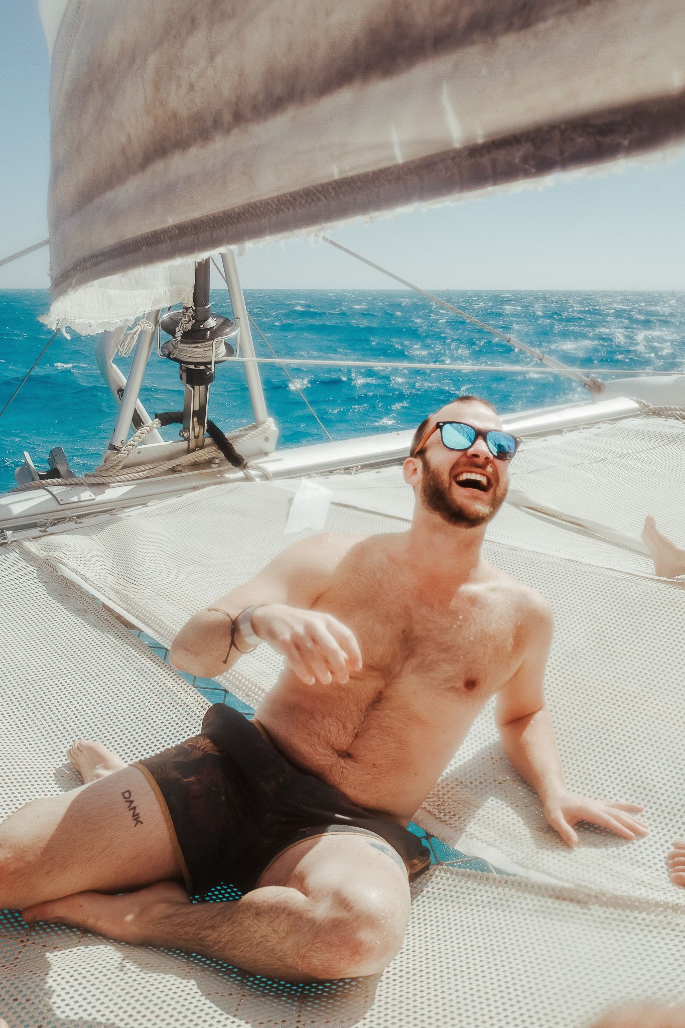 A shirtless person laughing while relaxing on a catamaran sail boat deck in bright turquoise waters.