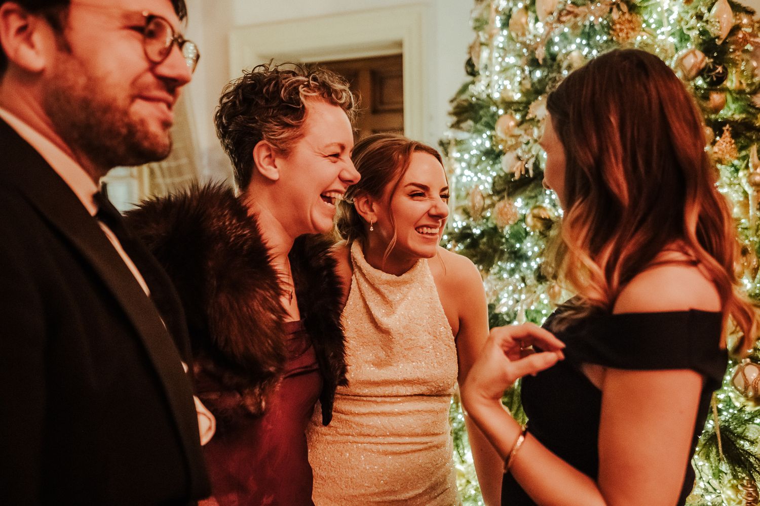 Guests sharing laughs and conversation near a decorated Christmas tree at an evening wedding celebration.