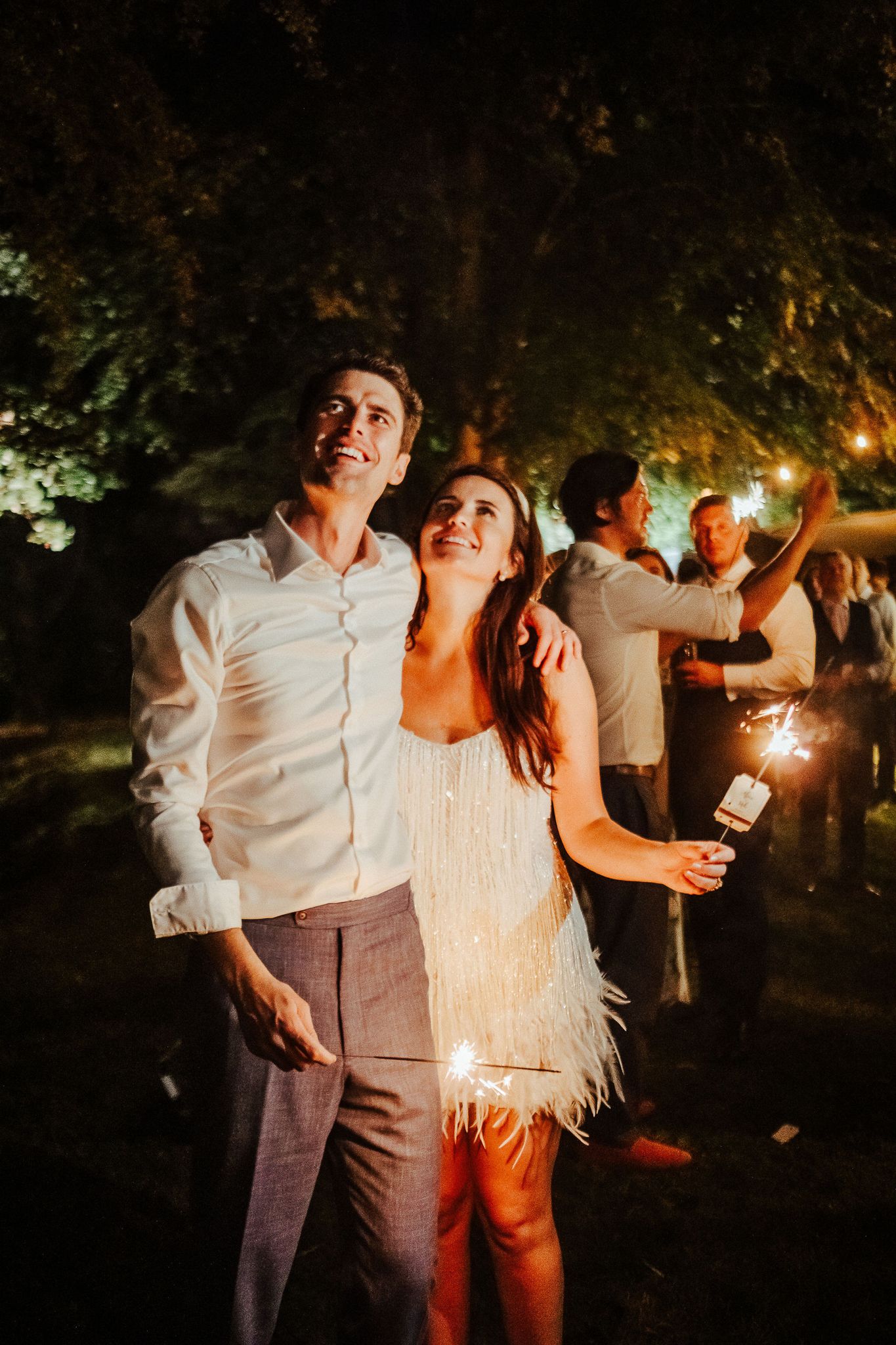 A couple celebrates at a nighttime wedding reception under trees with festive lighting.