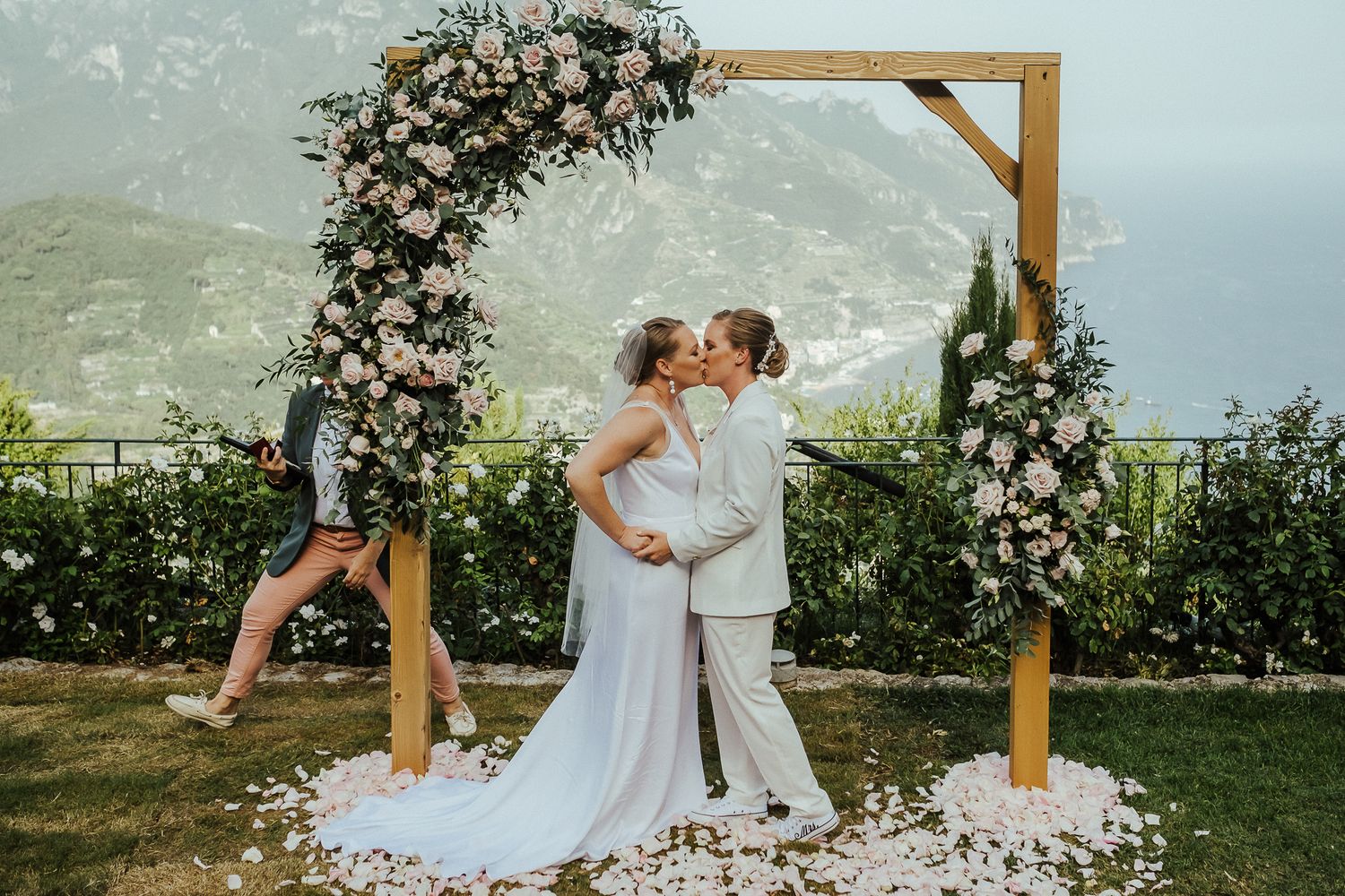 Newlyweds share their first kiss under a wooden arch decorated with white flowers at a mountaintop venue.