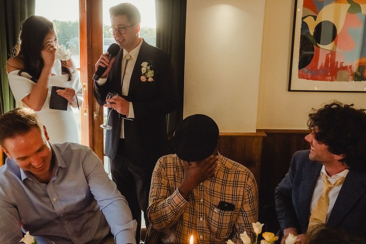 A group of people are gathered at a social event, seated at a table with drinks while others stand nearby.
