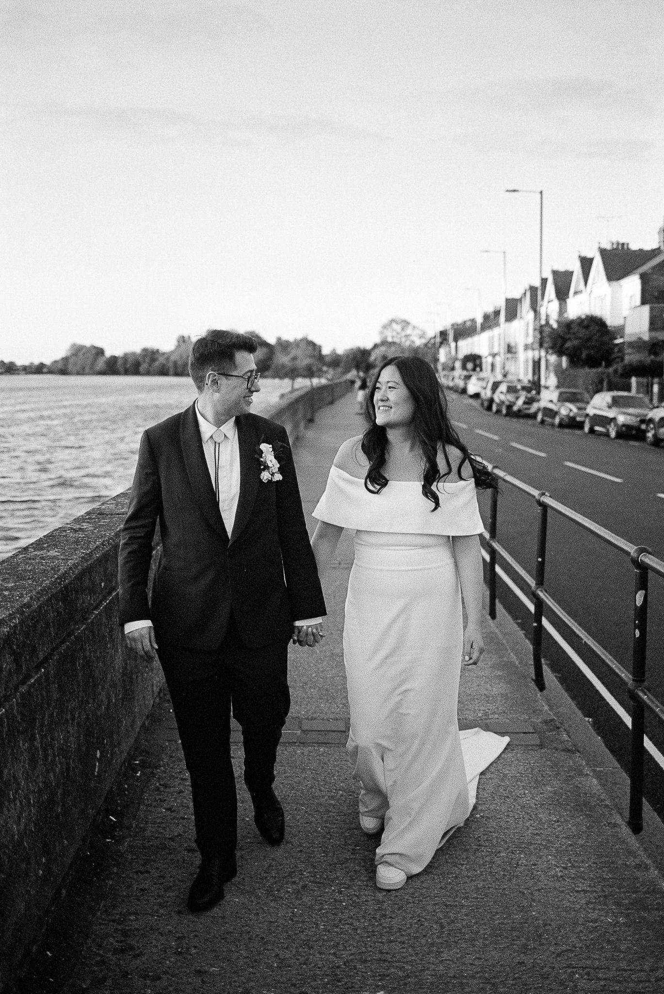 A couple in wedding attire walks happily along a waterfront promenade with row houses visible in the background.
