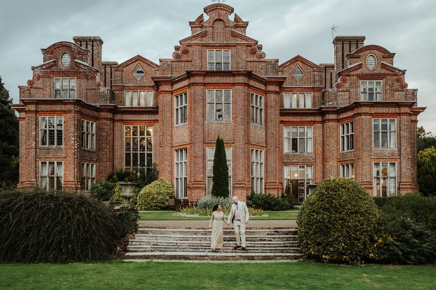 A grand red brick Victorian manor house with ornate Dutch-style gables and manicured gardens in the English countryside.