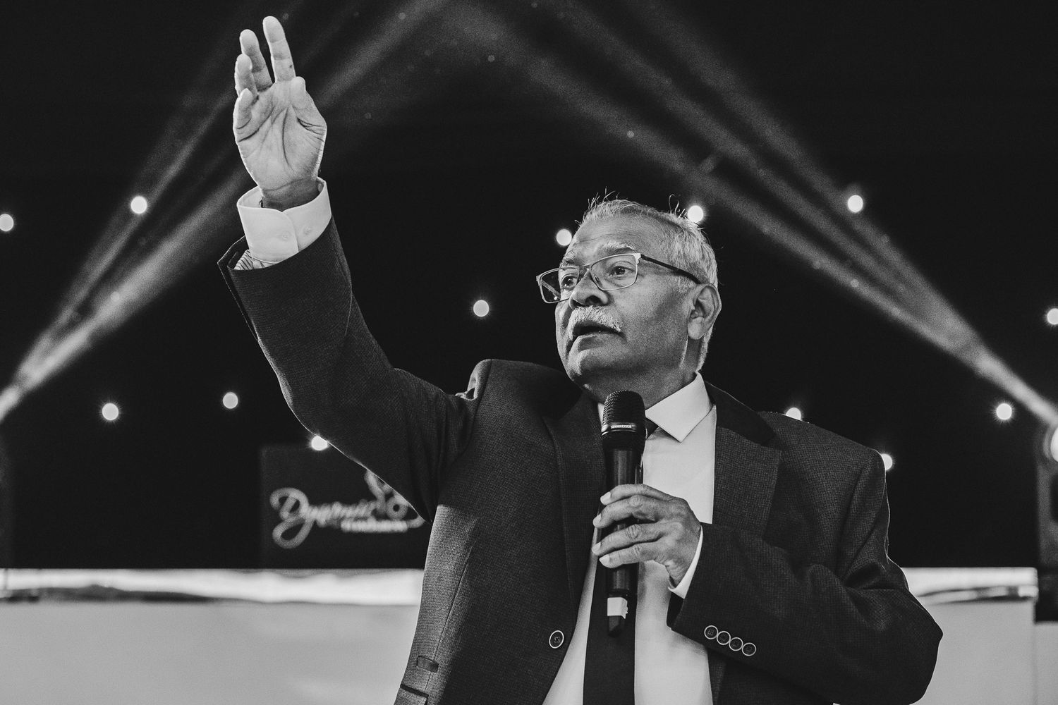 A person in a suit holding a microphone on stage with dramatic lighting beams in the background.