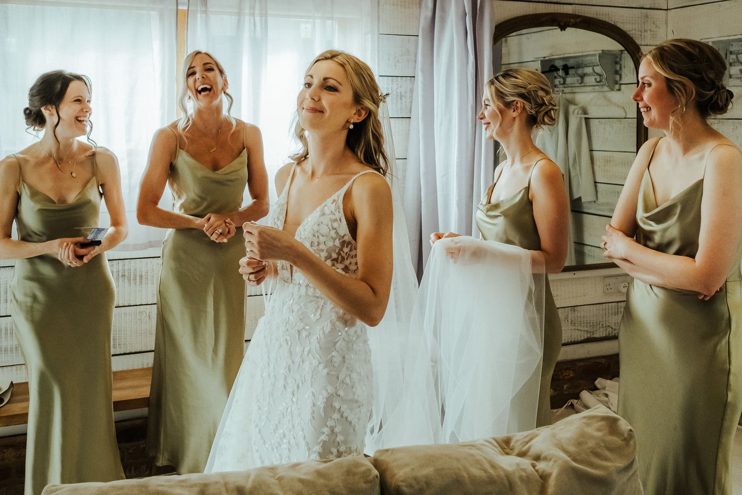 A group in sage green dresses prepares for a wedding celebration in a bright room.