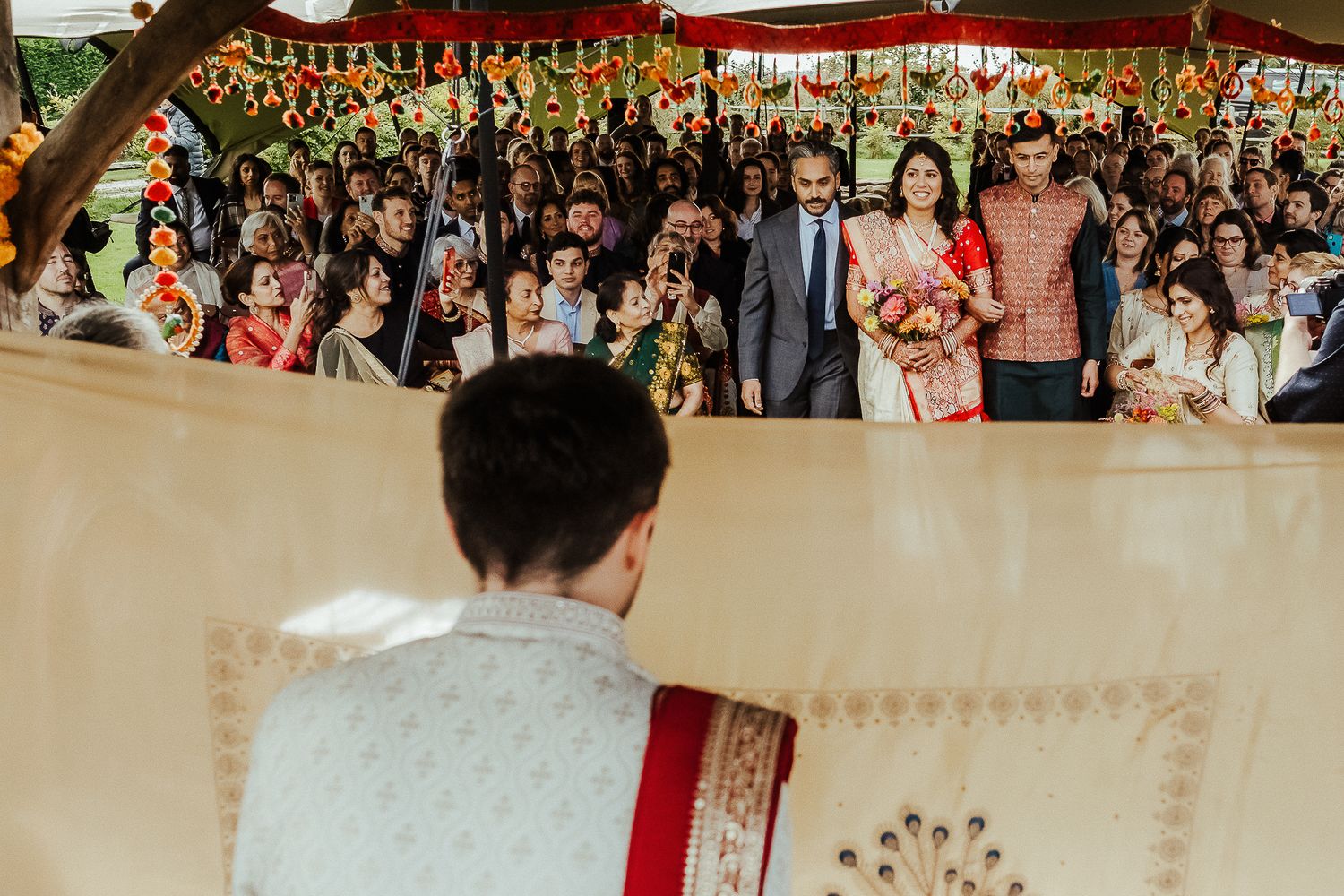 Wedding guests gather under a red canopy watching a traditional ceremony unfold at an outdoor celebration.