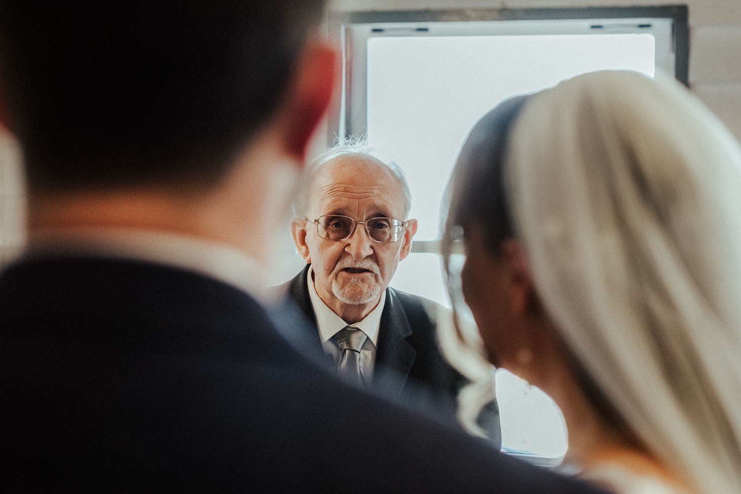 An older officiant looks on during an intimate wedding ceremony indoors.
