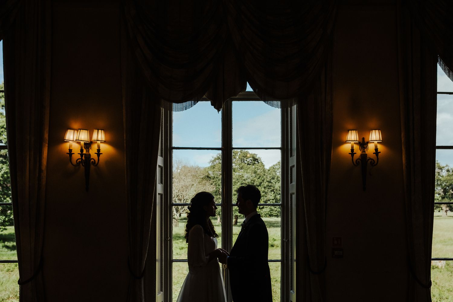 Silhouetted couple stands in front of bright window with ornate wall sconces creating romantic ambiance.
