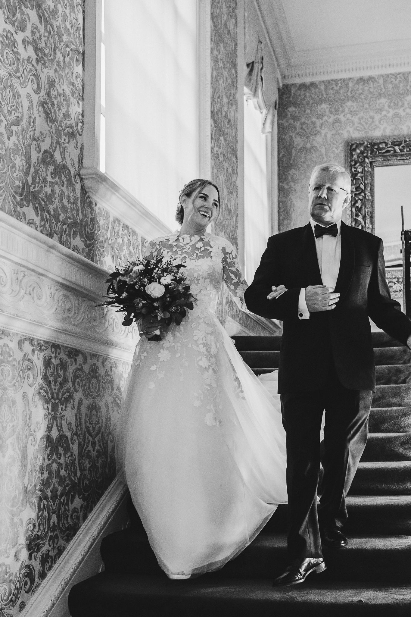Black and white photo of a bride descending ornate stairs in a vintage-style venue.