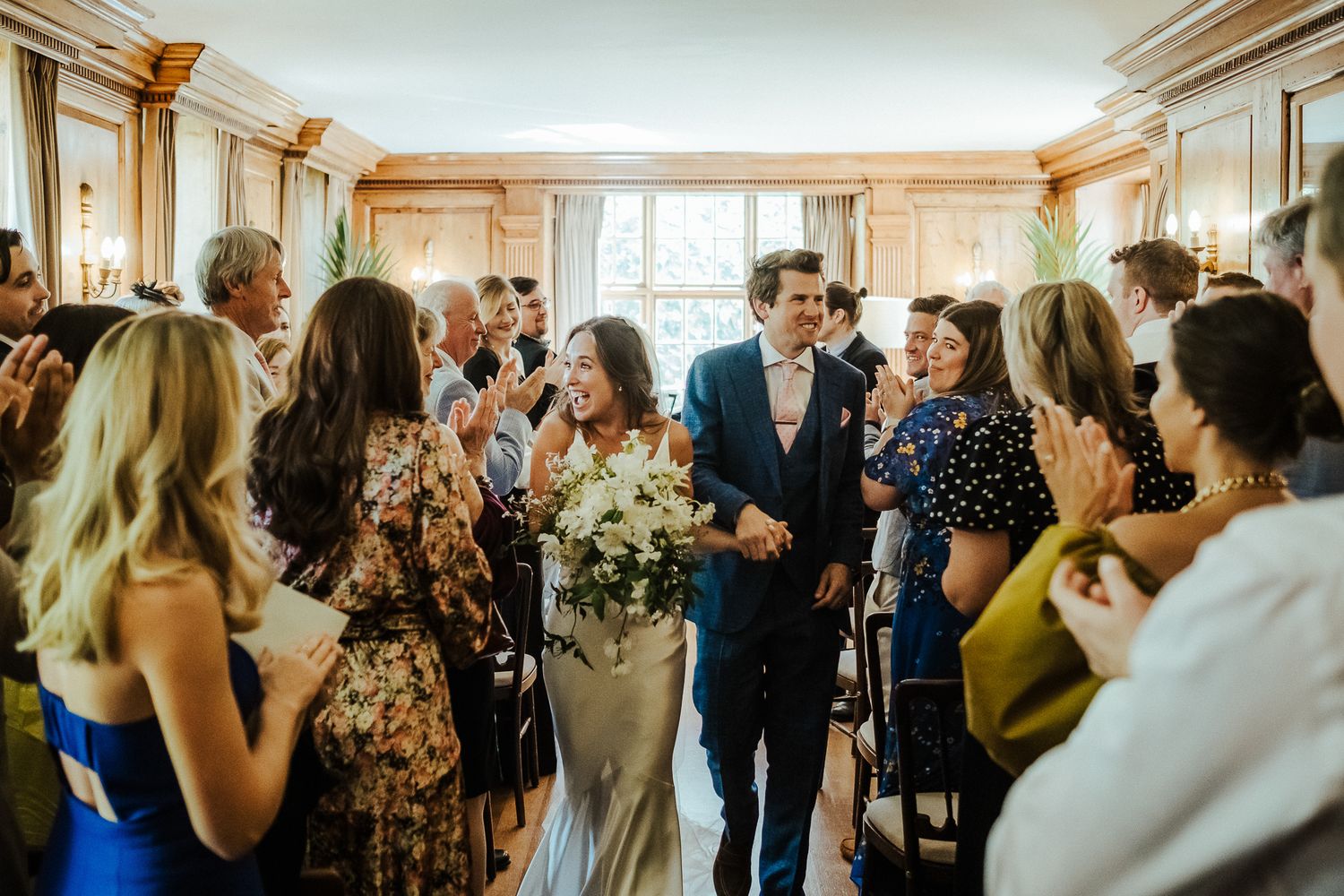 Guests surround the happy couple during a joyous wedding reception in a sun-filled room.