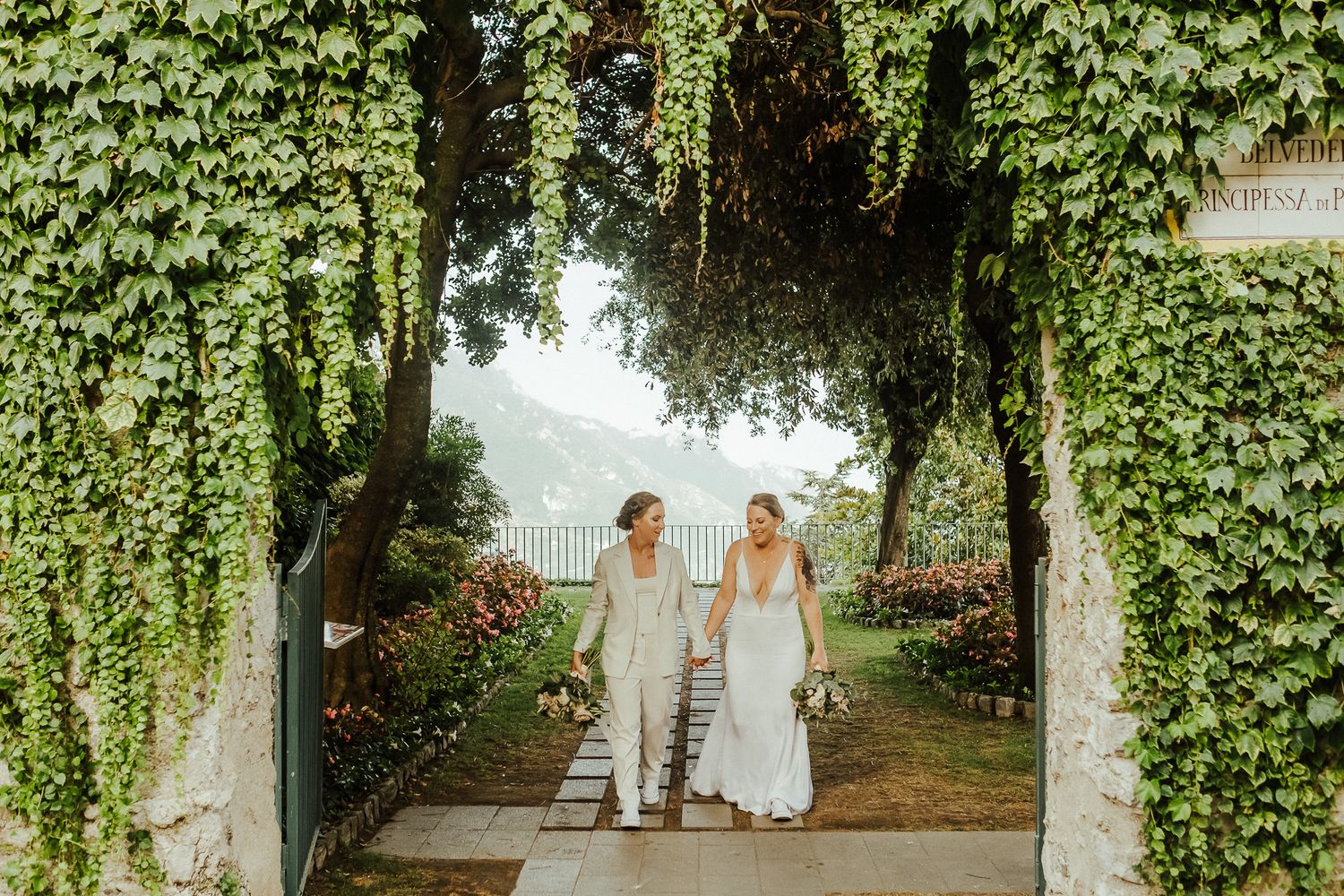 Couple walks through ivy-covered archway in elegant garden setting with mountain views.