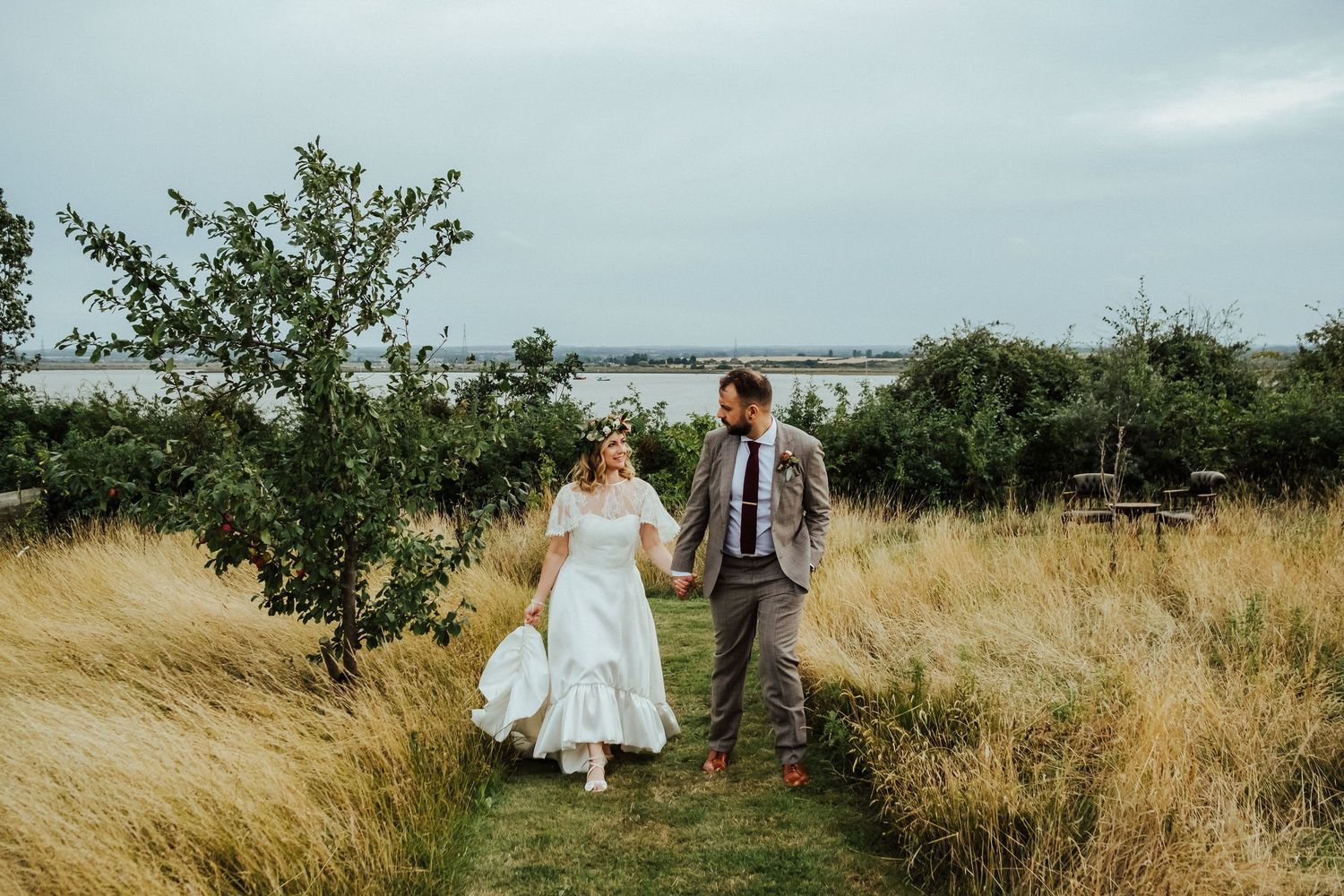 Wedding couple walking through tall grass field with rustic landscape backdrop.