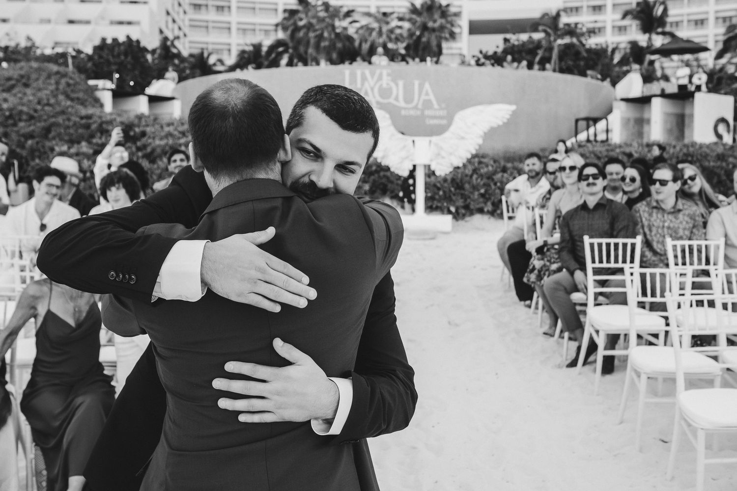 Two people share an emotional embrace during an outdoor wedding ceremony with white chairs.