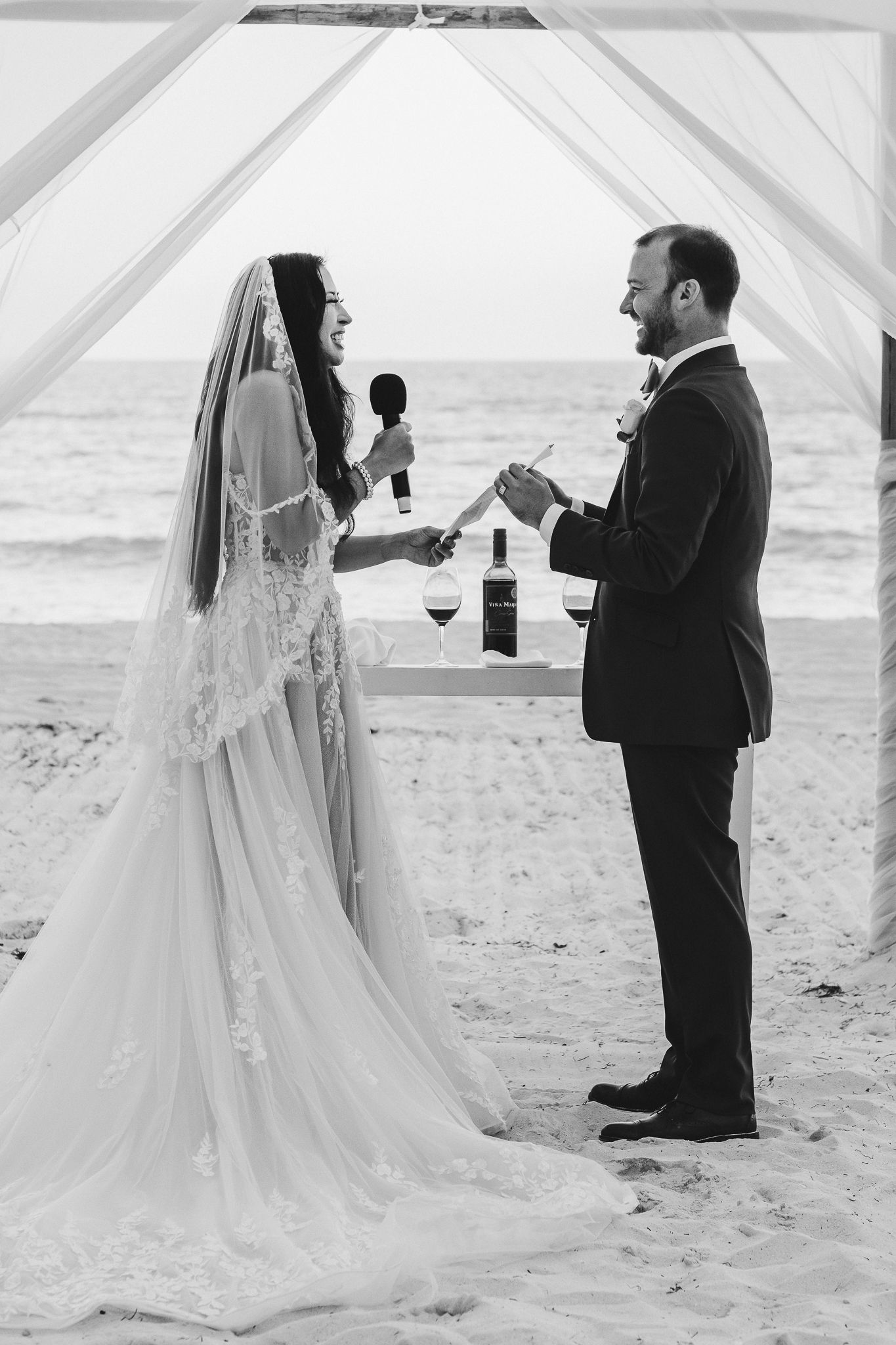 Black and white beach wedding photo of couple exchanging vows under wooden arch.