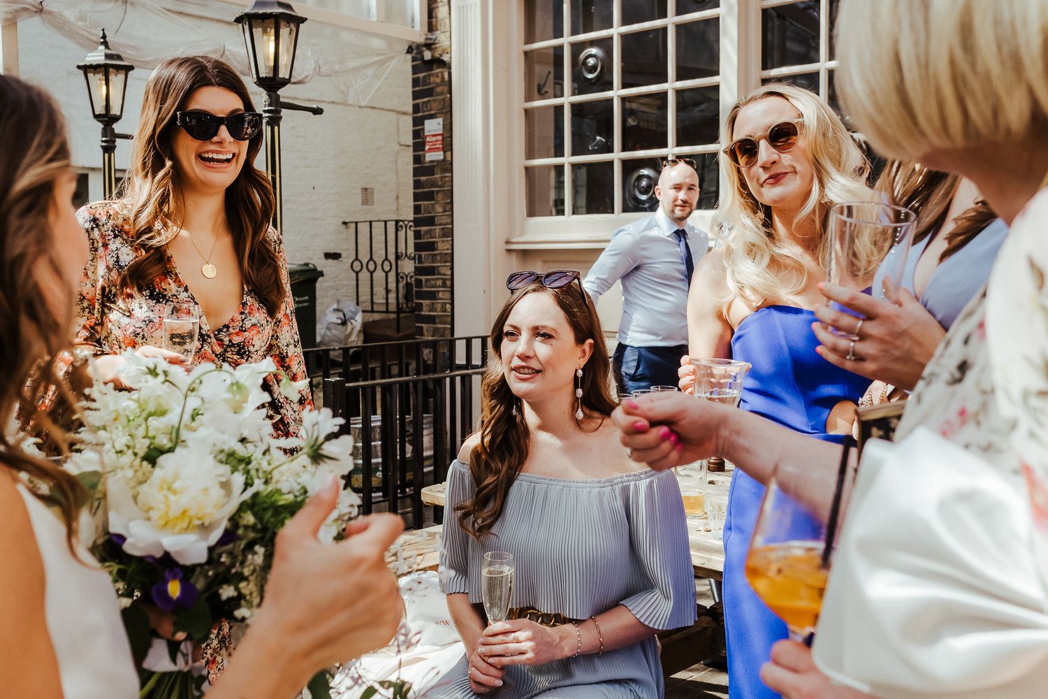 Wedding guests gather on an outdoor patio with drinks and flowers during a celebration.