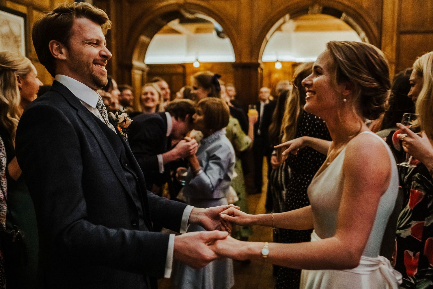 Wedding couple laughs joyfully while dancing together surrounded by guests in a historic venue.
