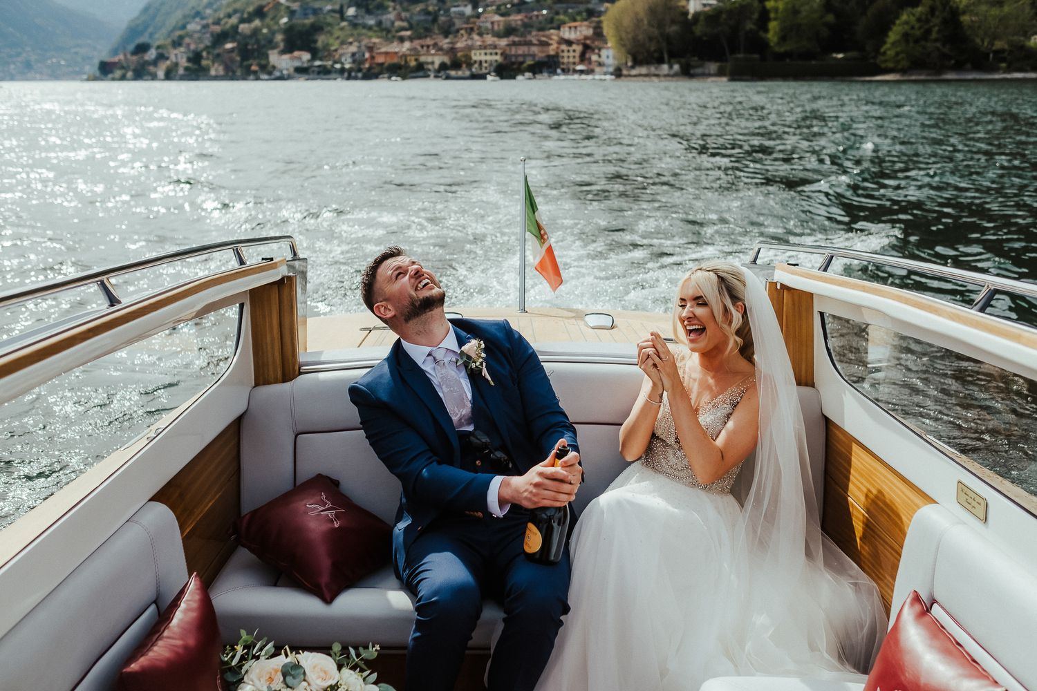 Couple laughing together on vintage wooden boat with Italian mountains in background.