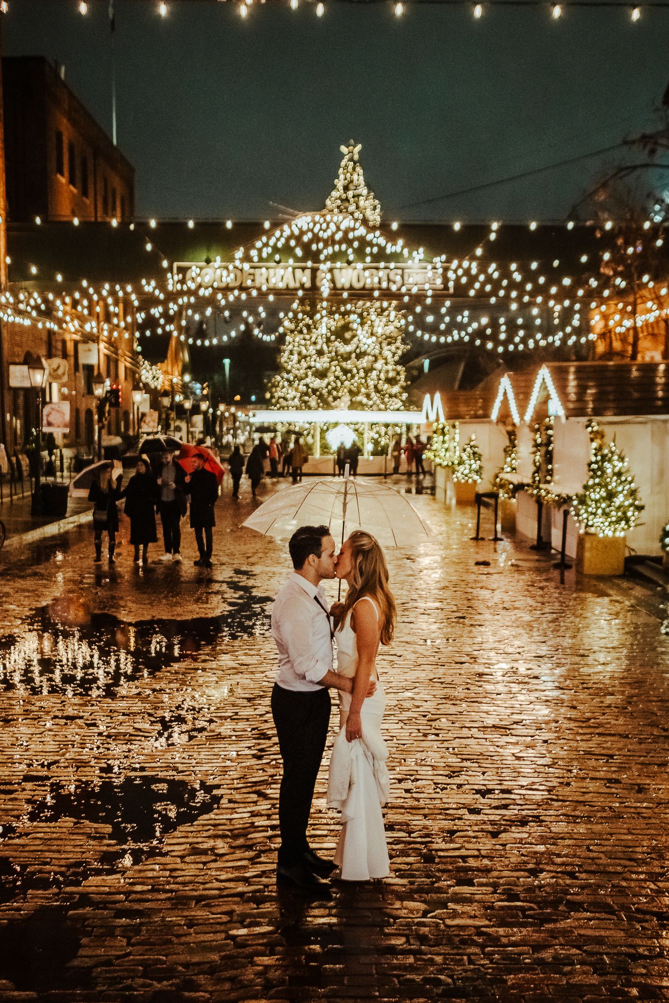 Romantic couple kisses on cobblestone street with holiday lights and Christmas decorations glowing above.