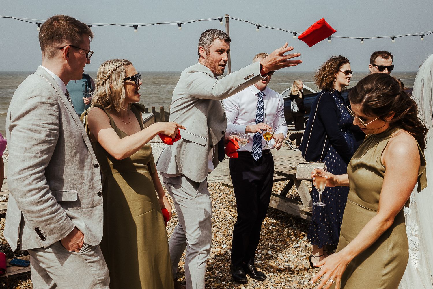Wedding guests in formal attire gather outdoors for drinks under string lights.