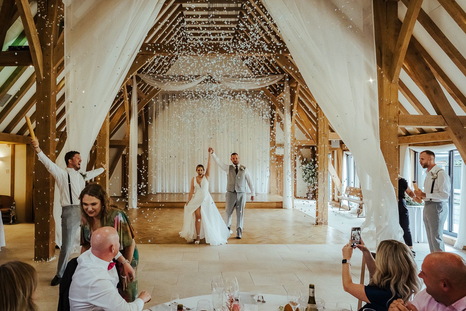 Rustic barn wedding ceremony with guests watching couple under wooden beams and hanging lights.