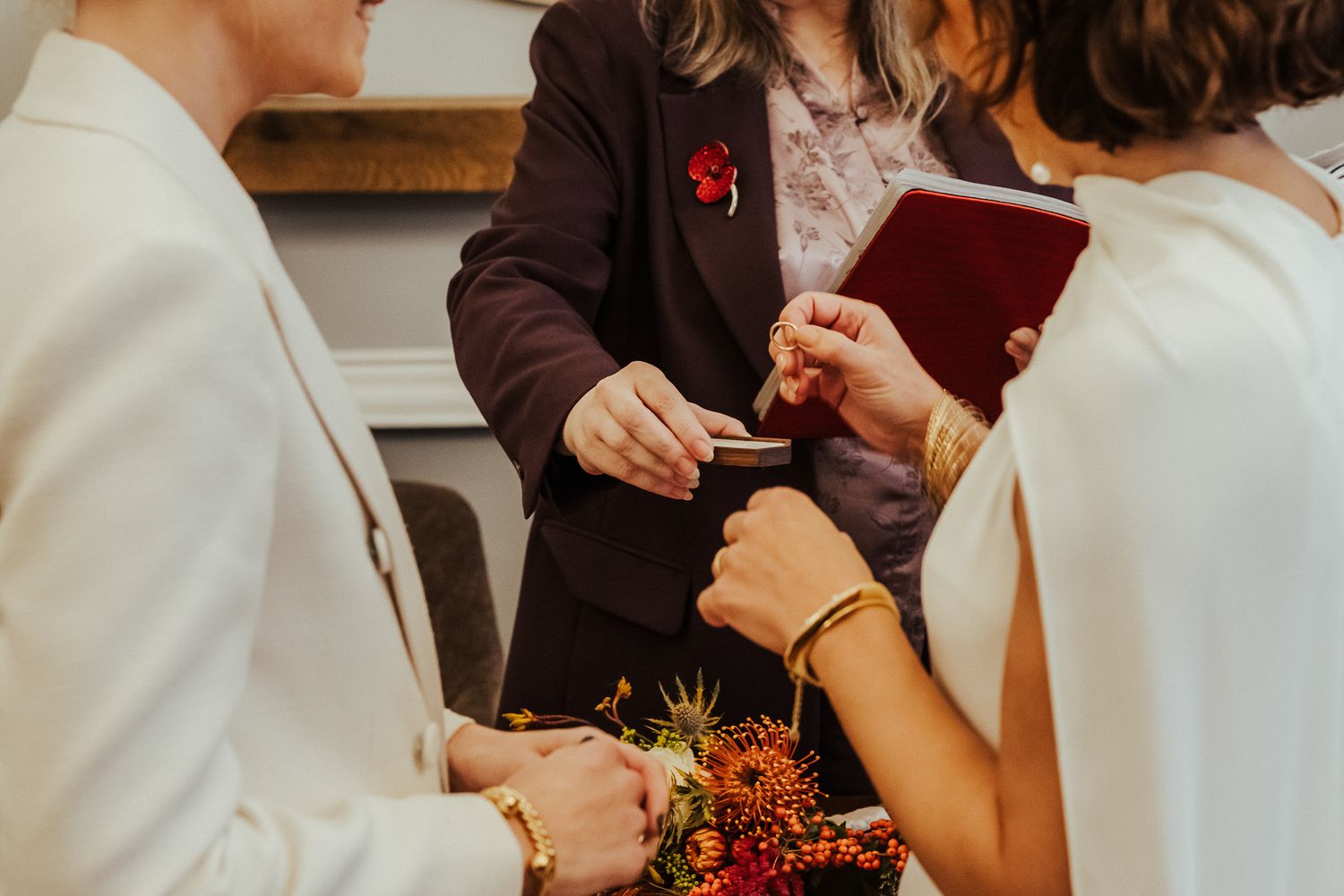 Wedding officiant in burgundy jacket holds red book while couple exchanges rings during intimate ceremony.