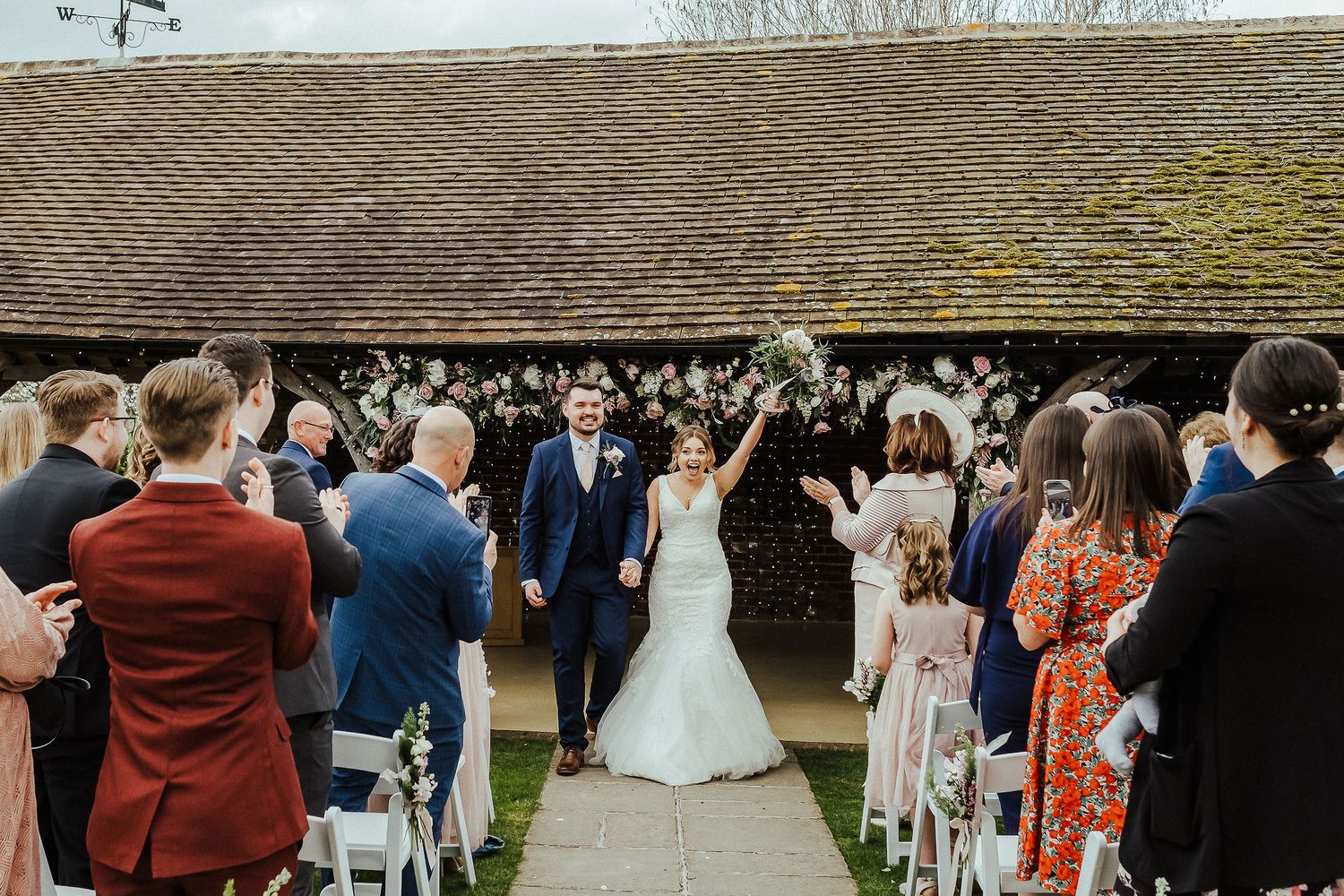Wedding guests throw confetti over newlyweds outside rustic venue.