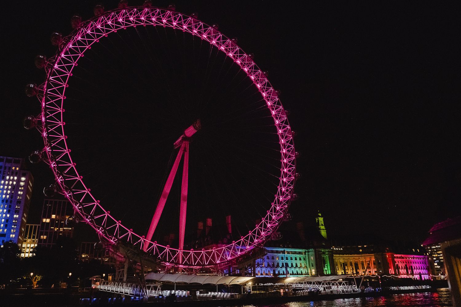The London Eye Ferris wheel illuminated in bright pink against the night sky along the River Thames.