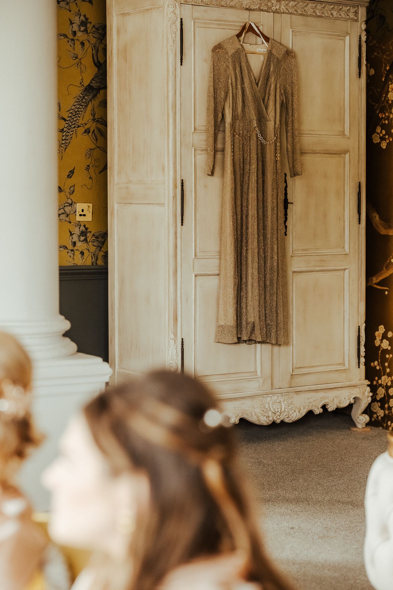 A long beige sparkly dress hangs on a white vintage armoire with floral wallpaper in the background.