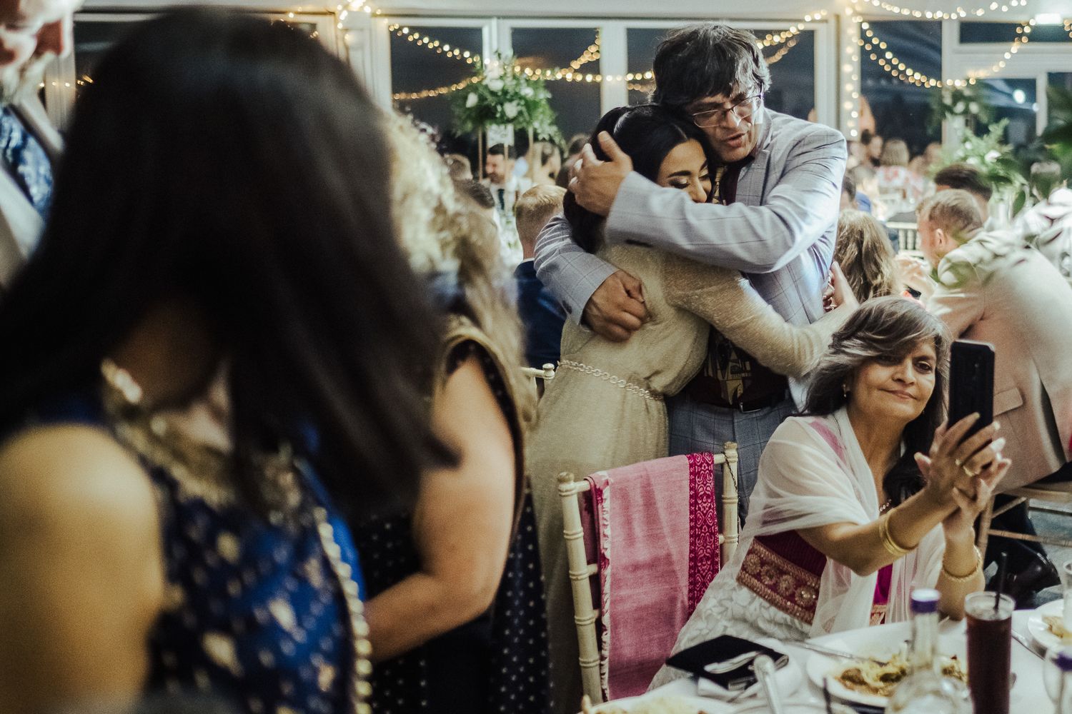 Wedding guests gather at a celebration, clinking glasses and toasting during an indoor reception party.