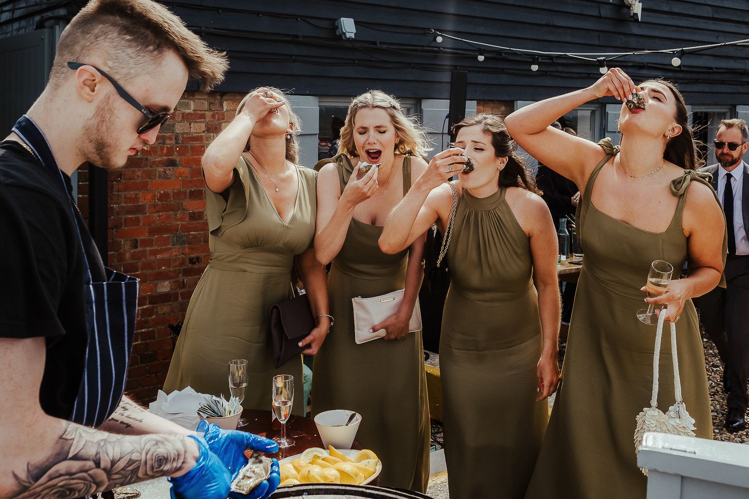Bridesmaids in olive green dresses react excitedly while someone pours drinks.