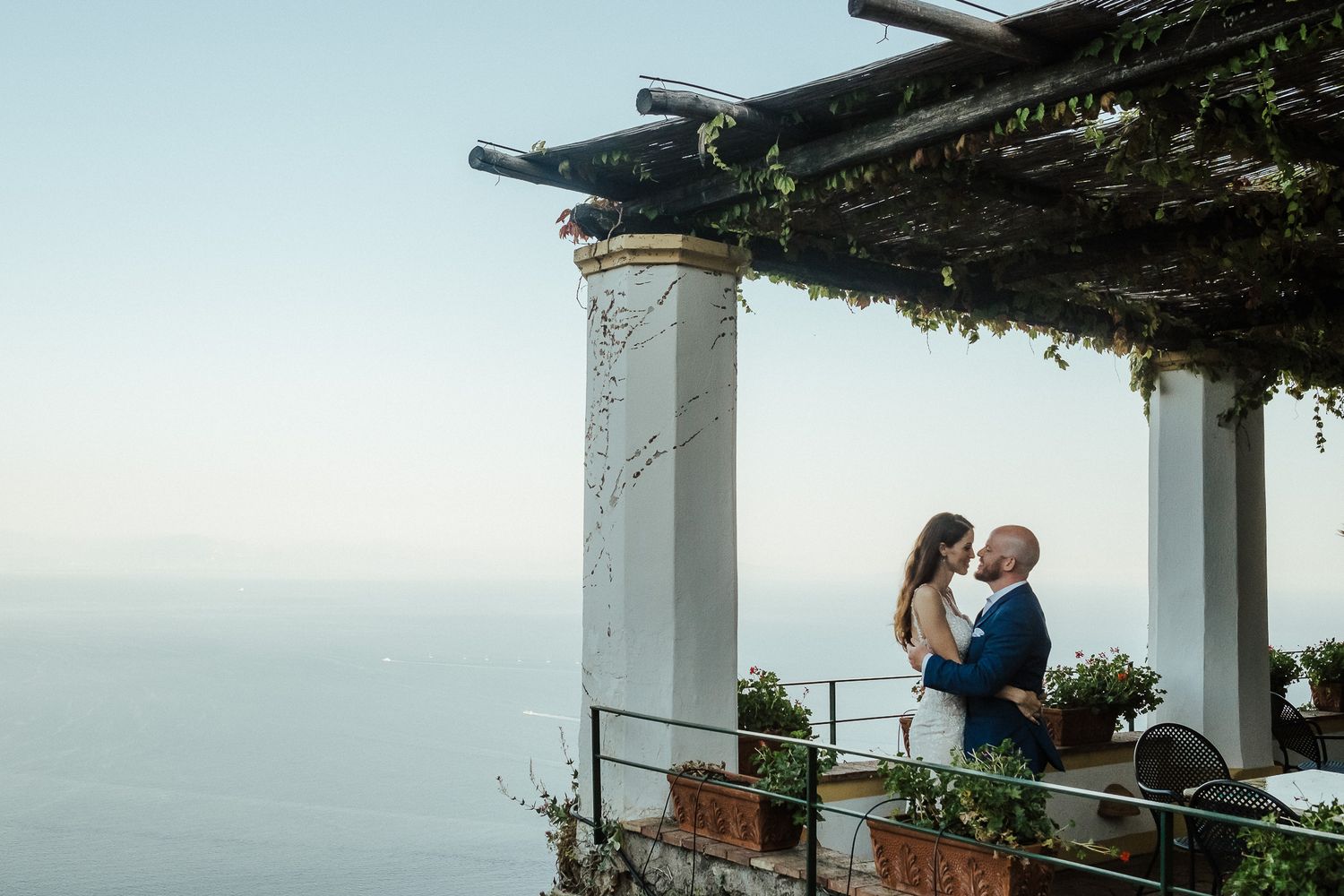 A couple embraces on a Mediterranean terrace overlooking the sea at sunset.