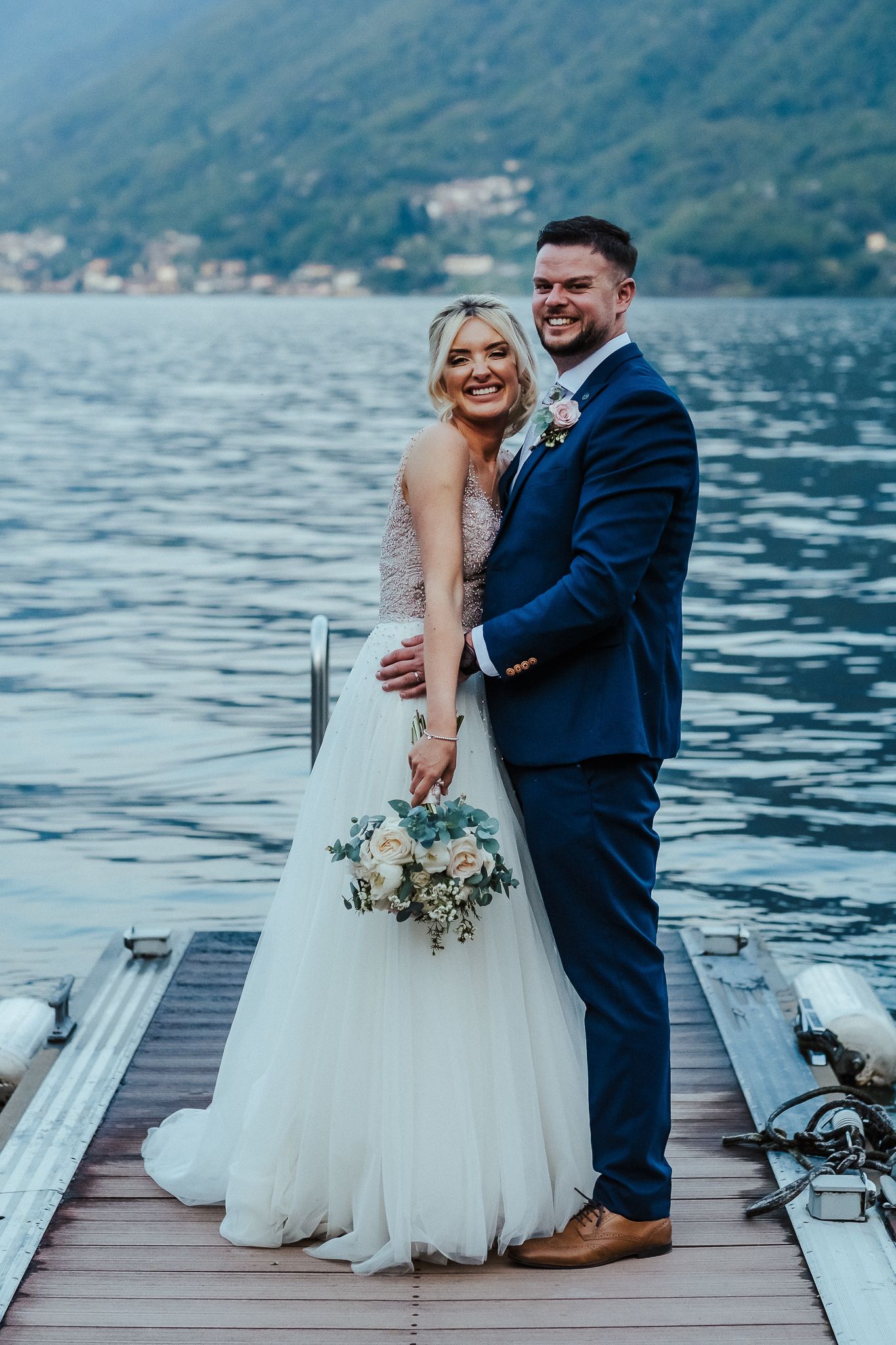 Bride in white gown holds bouquet while posing with groom in navy suit on wooden dock.