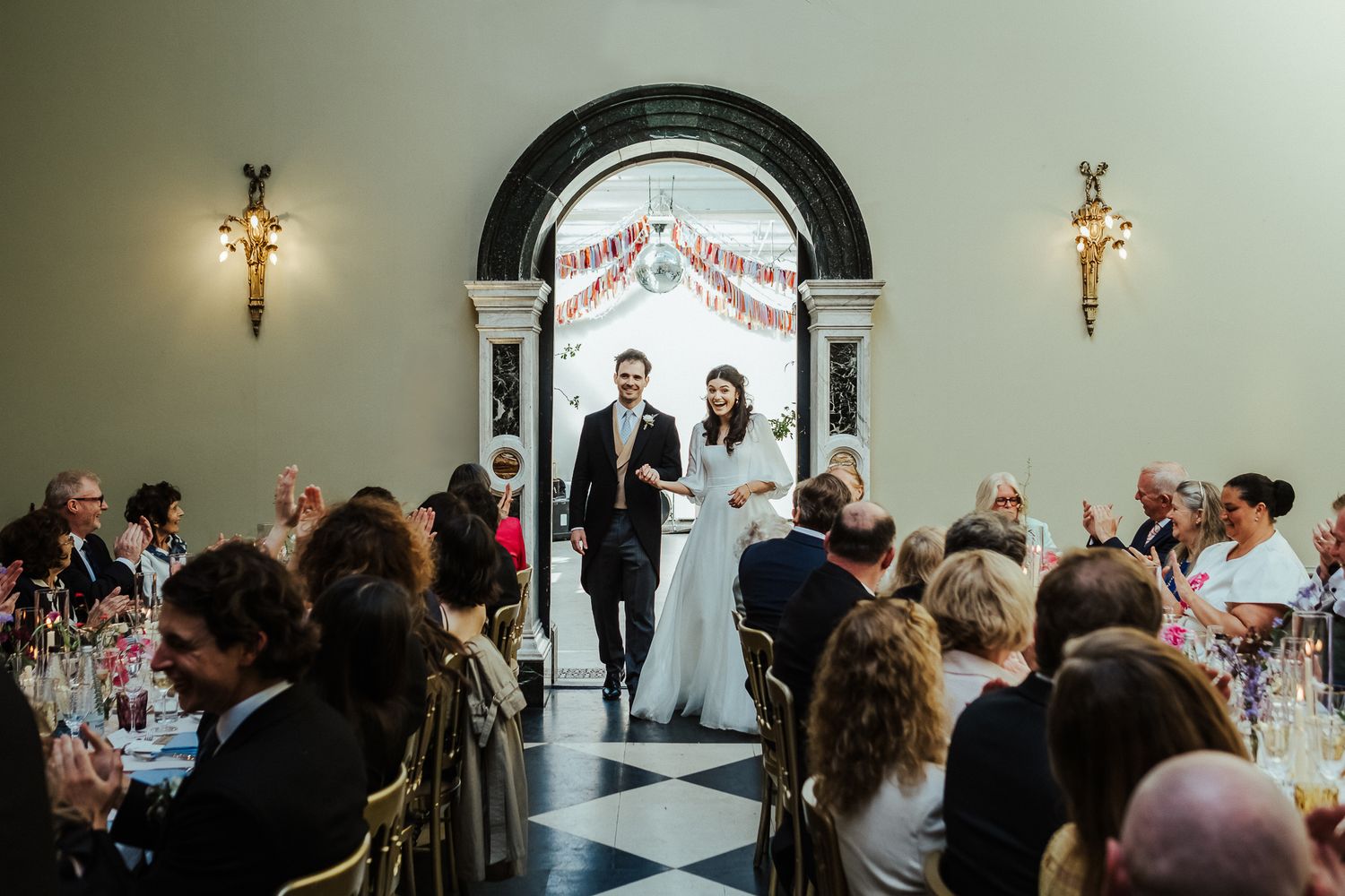 Wedding reception entrance with couple framed by black archway as guests dine at decorated tables.