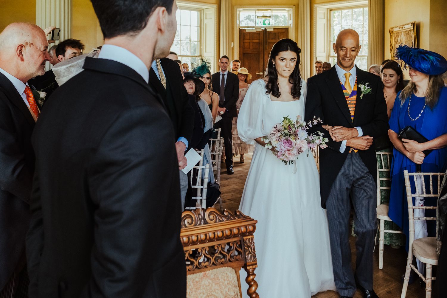 Wedding ceremony in a grand yellow room with elegant decor and guests seated in white chairs.