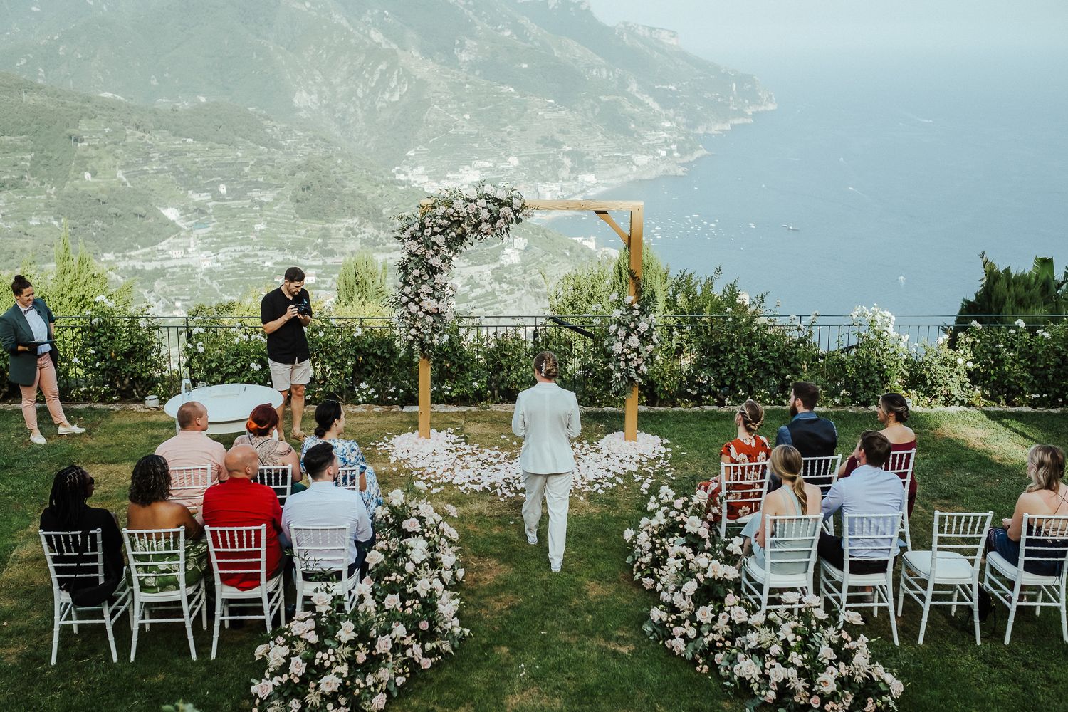 Intimate wedding ceremony setup with white chairs and floral arch overlooking coastal views.