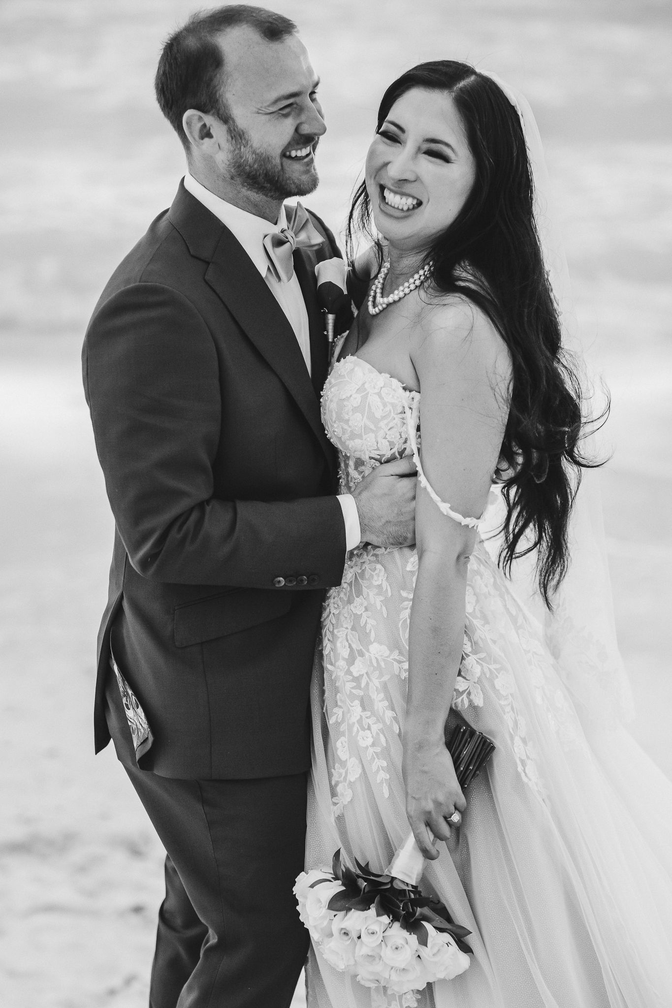 Black and white portrait of joyful newlyweds laughing together on beach.