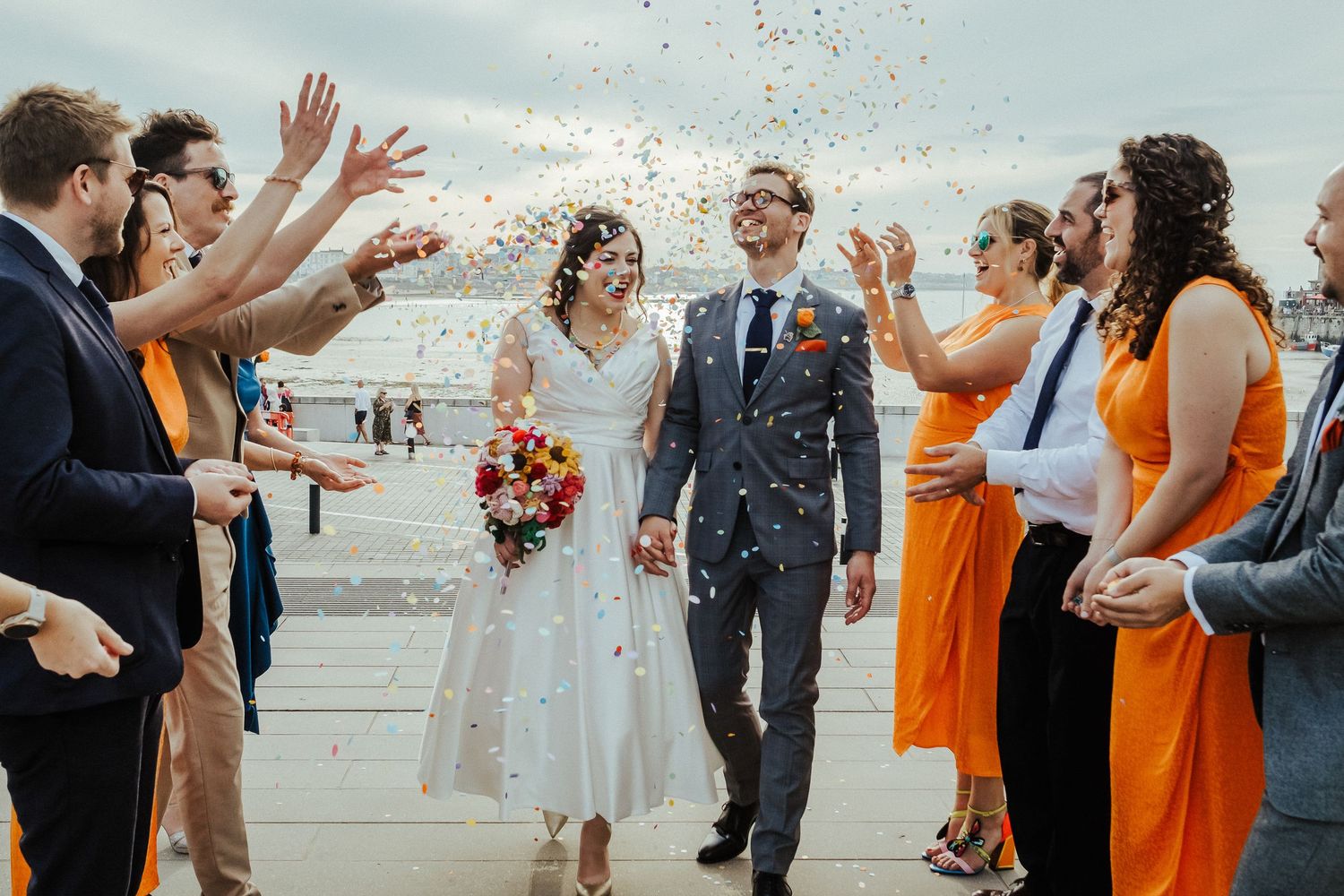 Wedding party celebrating with confetti on a beach boardwalk with guests in orange dresses.