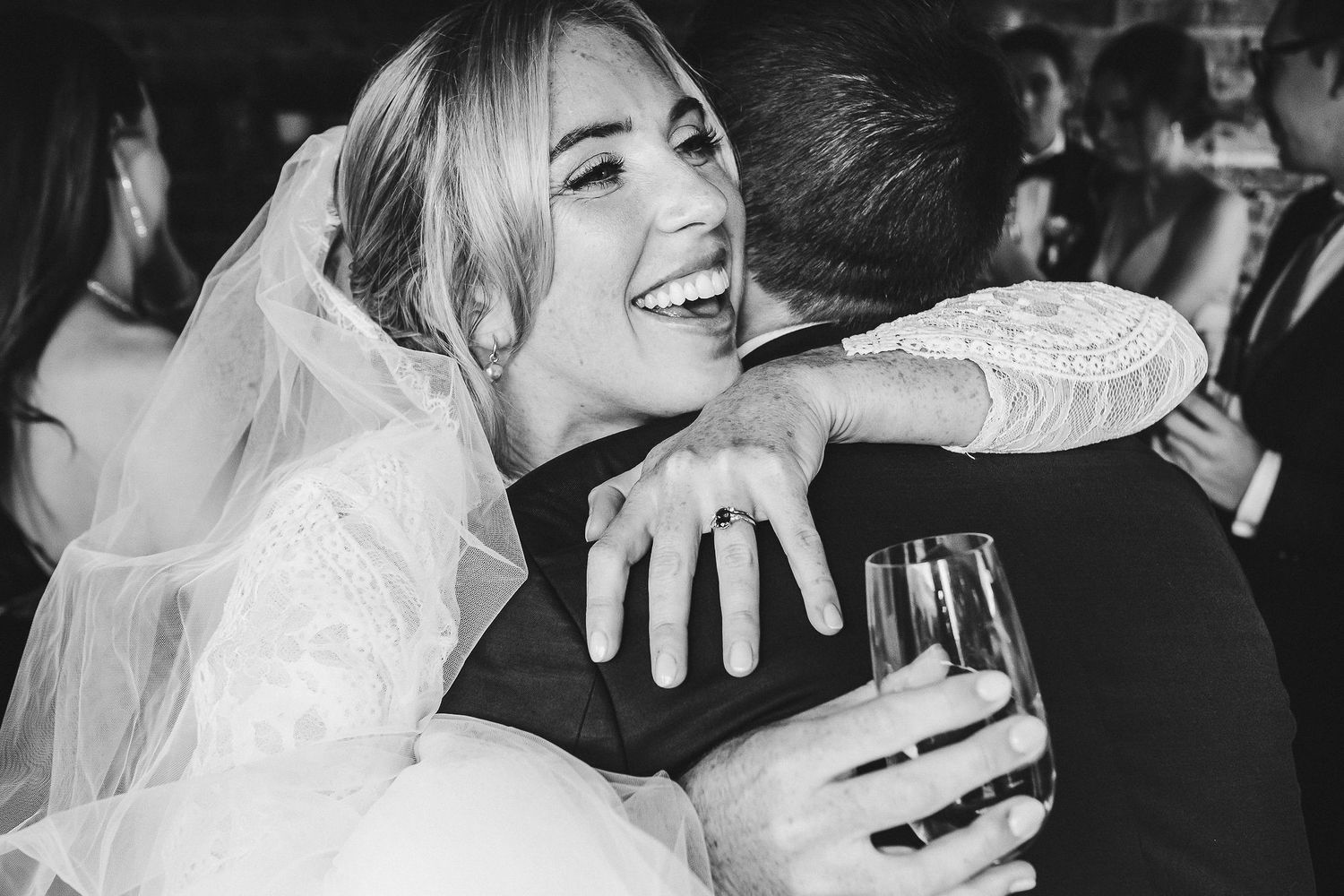 Black and white photo of bride hugging and laughing while holding champagne glass at reception.