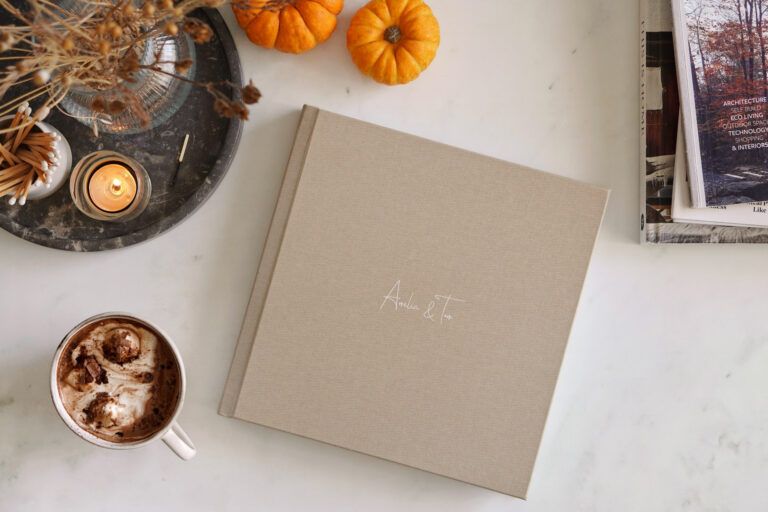 Flat lay of wedding album with autumn decorations and hot drink on white surface.