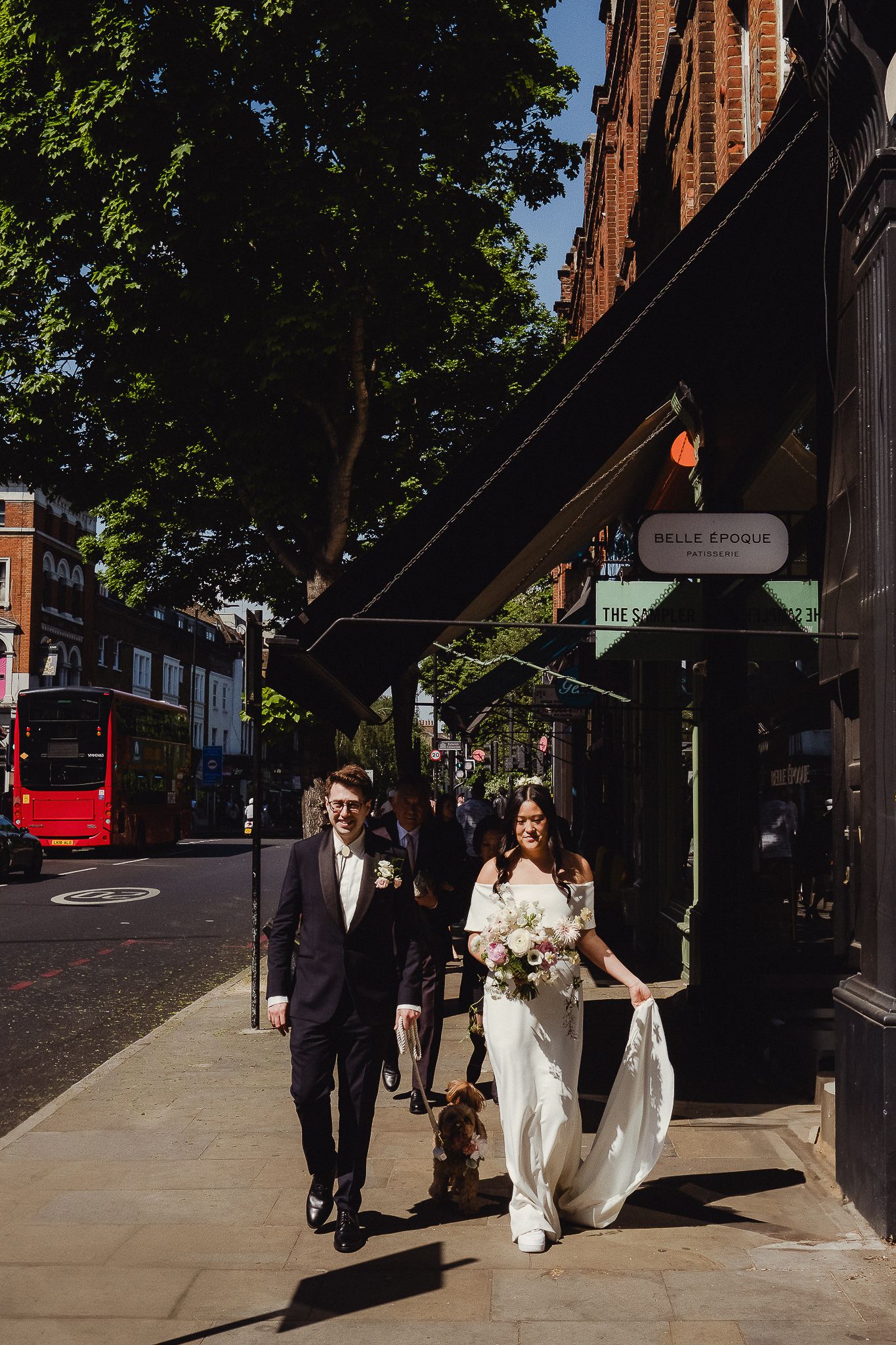 A couple walking together on a London sidewalk in sunlight with a red bus and brick buildings in the background.