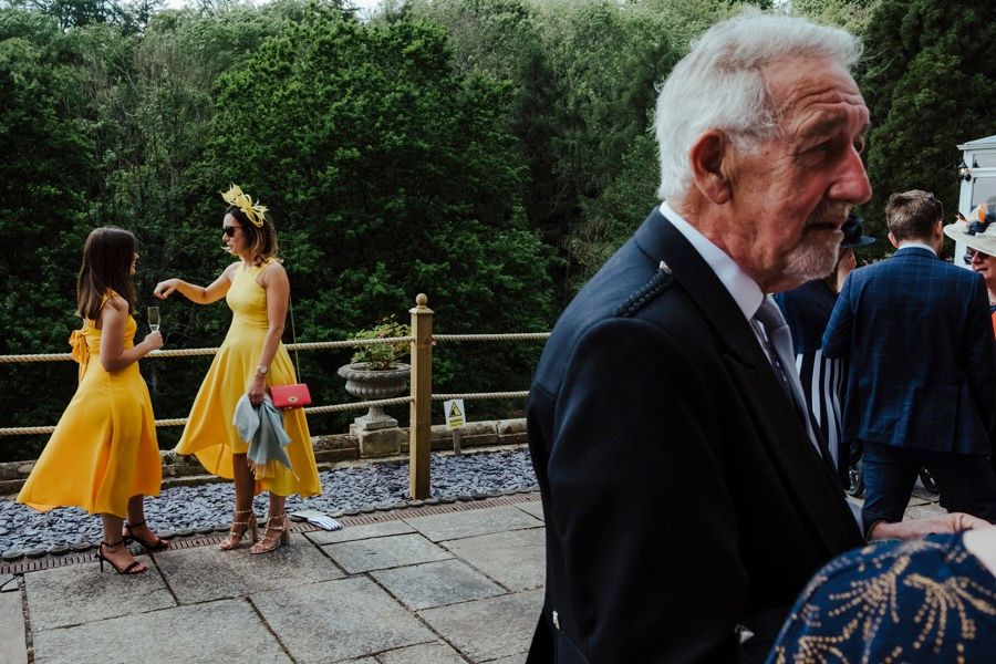 Candid outdoor wedding scene with yellow dresses and formal attire against natural backdrop.